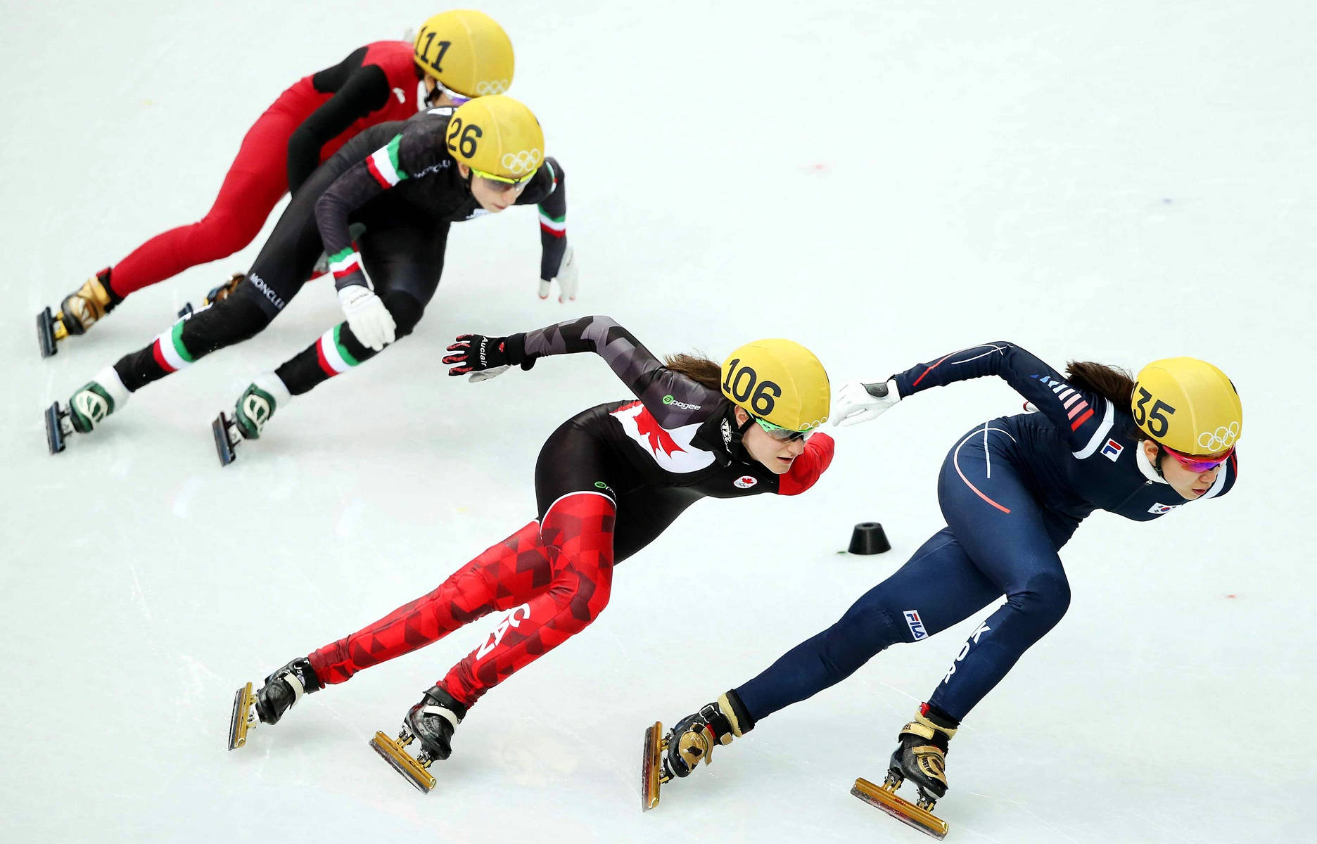 Olympics 桌布 壁紙 — 四位速度滑冰選手在激烈競爭中，在室內溜冰場的彎道上傾身，穿著國家隊制服和黃色頭盔。