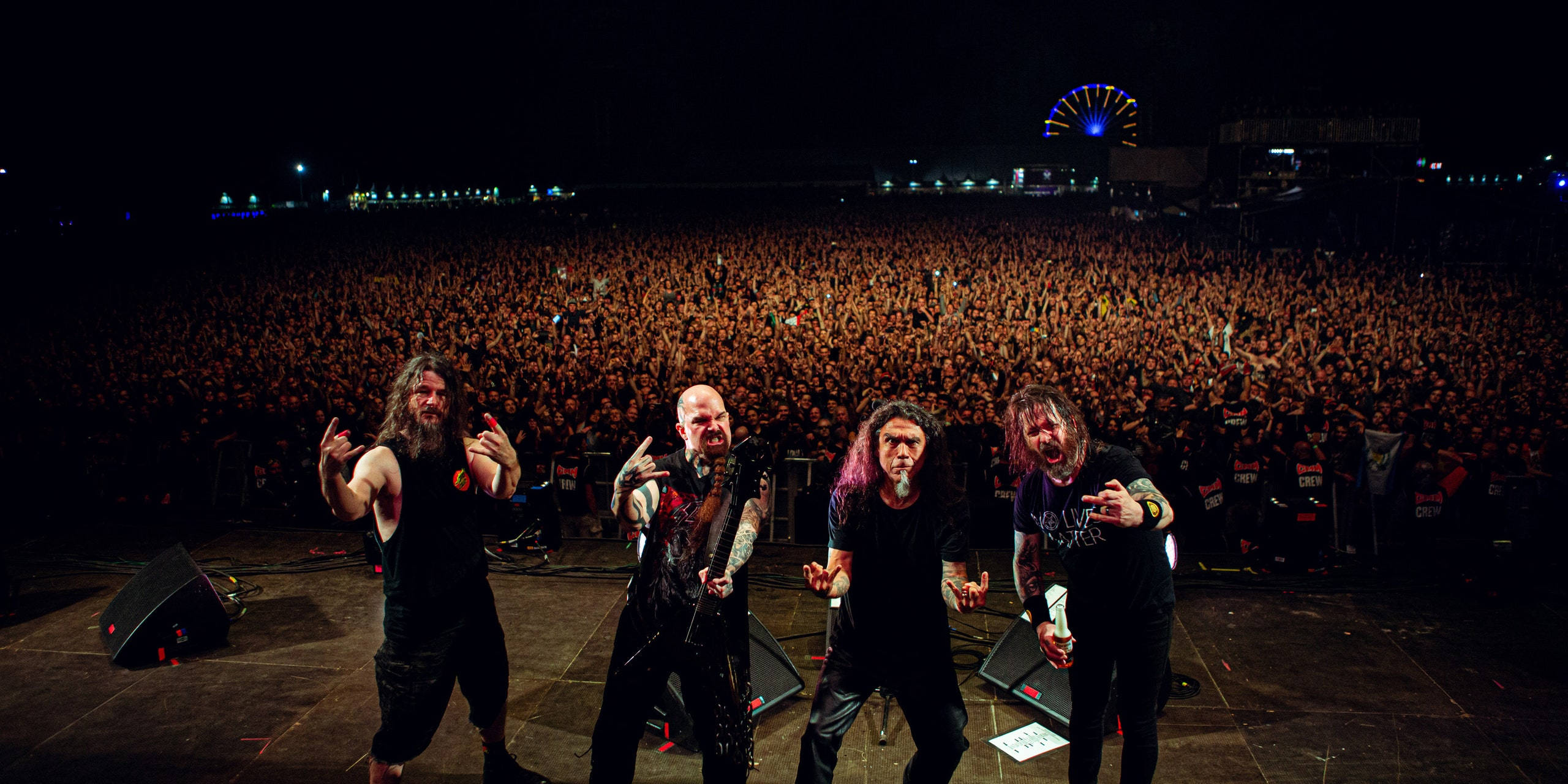 Slayer 桌布 壁紙 — 四位重金屬樂隊成員在夜空下，遠處摩天輪燈光中，在舞台上充滿活力地為大量觀眾表演。