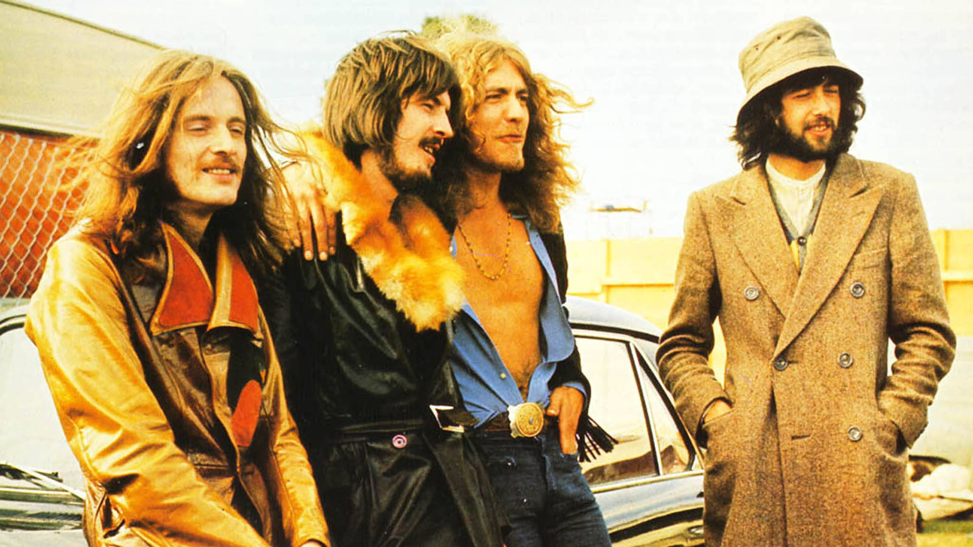 band 桌布 壁紙 — 四位長髮男士穿著1970年代時尚，隨意地站在一輛復古車旁，背景有鐵絲網圍欄。