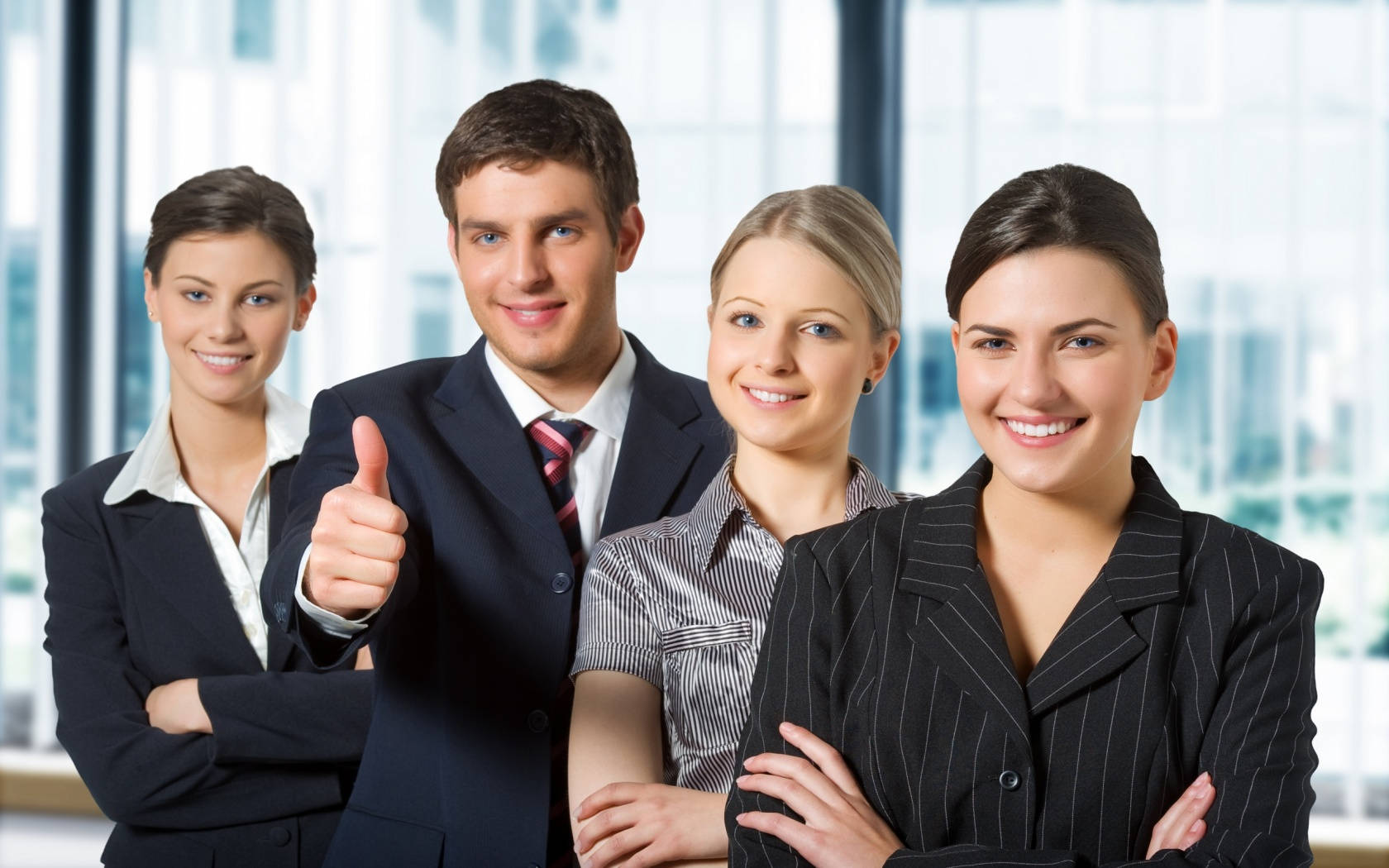 Teamwork 桌布 壁紙 — 四位面帶微笑的商務專業人士身穿西裝，在明亮的辦公室窗戶背景下自信地豎起大拇指擺姿勢。