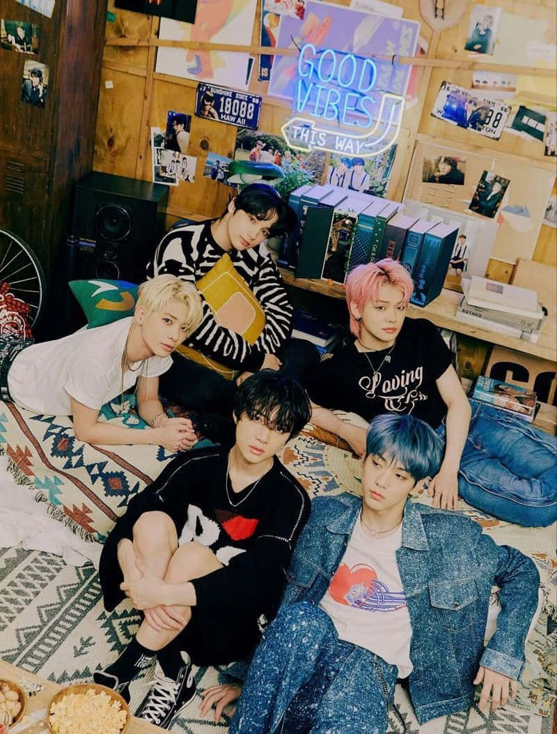 K-pop 桌布 壁紙 — 四位頭髮色彩繽紛的年輕男子在一個舒適的木質牆面房間裡，隨意地躺在圖案地毯上，房間裡佈滿了海報和霓虹燈牌。
