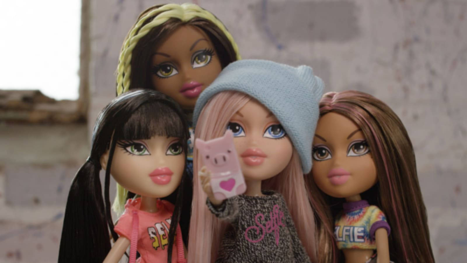 Bratz 桌布 壁紙 — 四個 Bratz 娃娃一起擺姿勢，其中一個在風化的磚牆前拿著一個豬主題的電話。