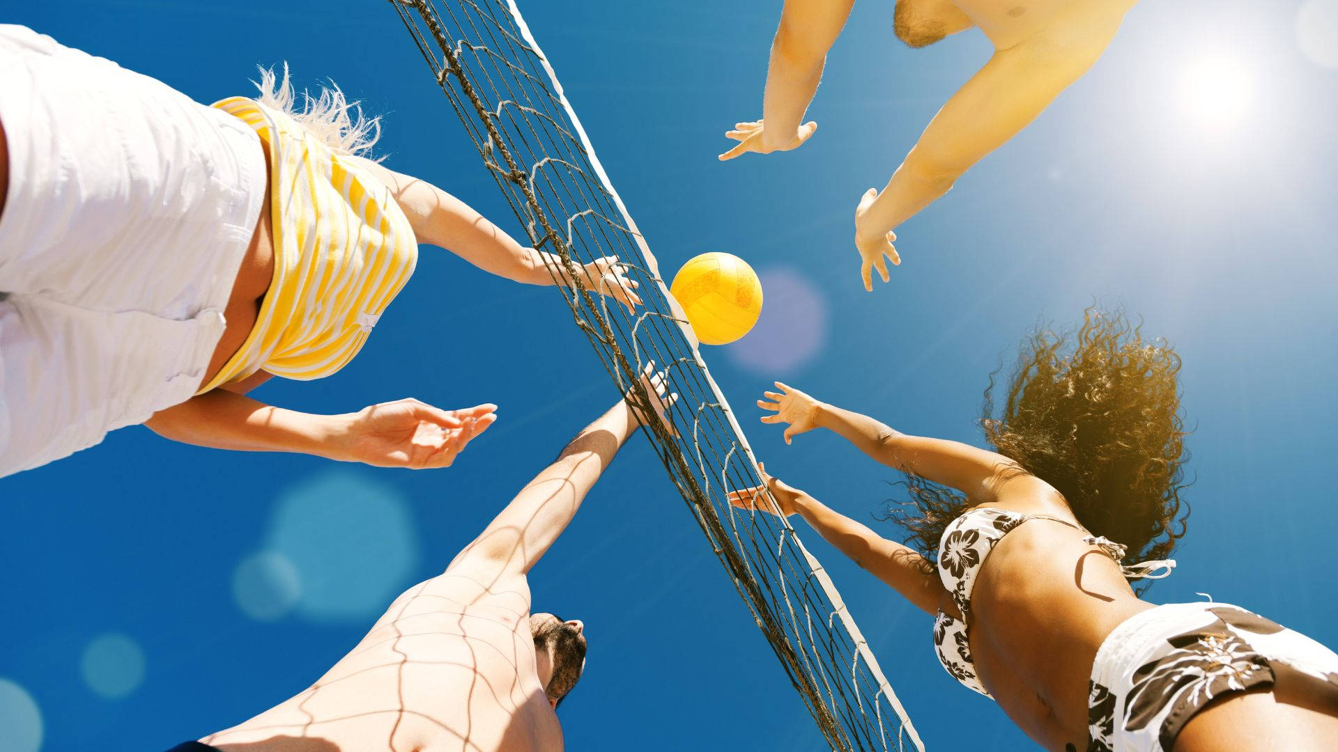 Volleyball Aesthetic 桌布 壁紙 — 四個人在燦爛陽光下打沙灘排球，黃色球在空中，背景是鮮豔的藍天。