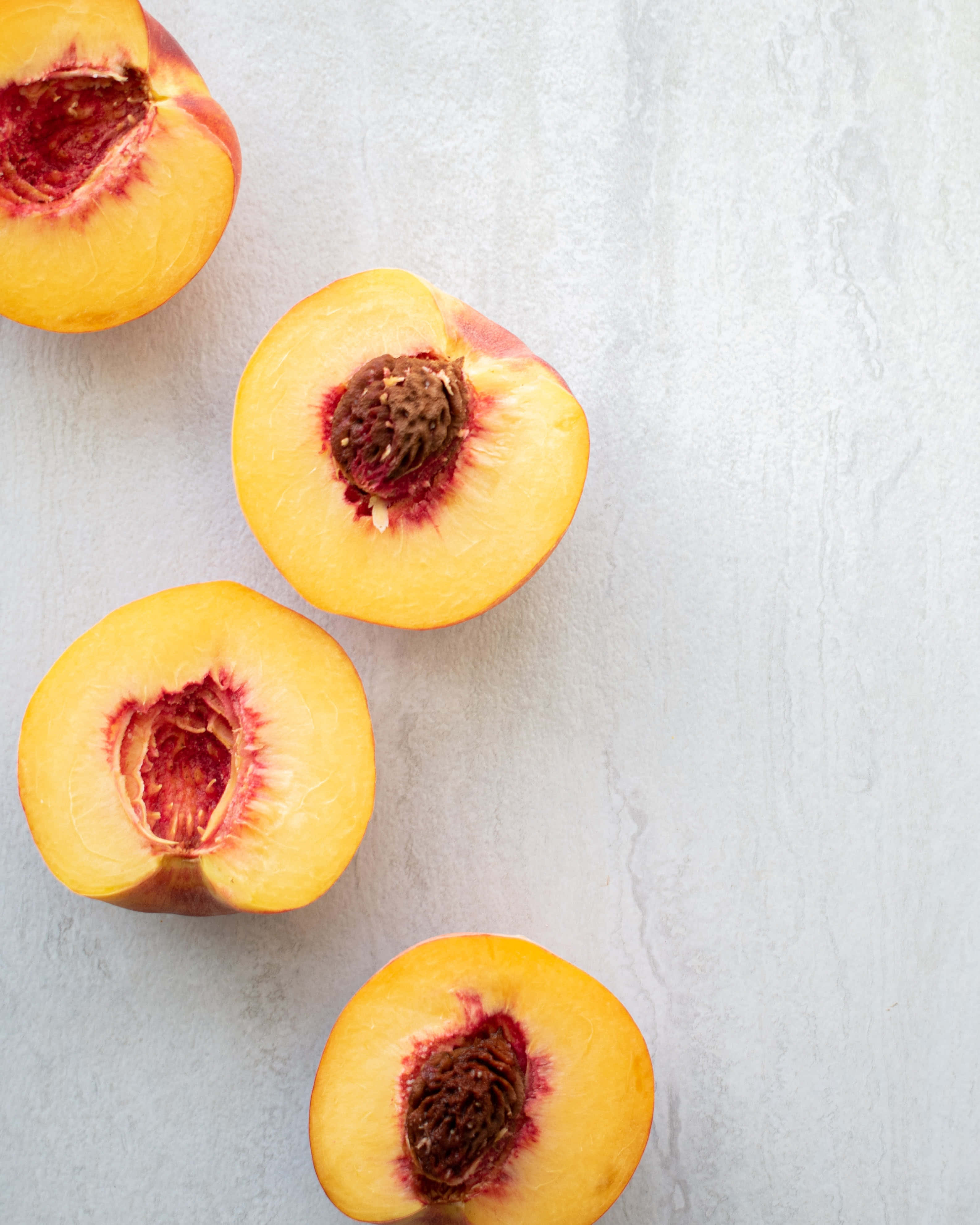 Peaches 桌布 壁紙 — 四個切半的桃子，多汁的黃色果肉帶有紅色暈彩，放在淺色紋理表面上。