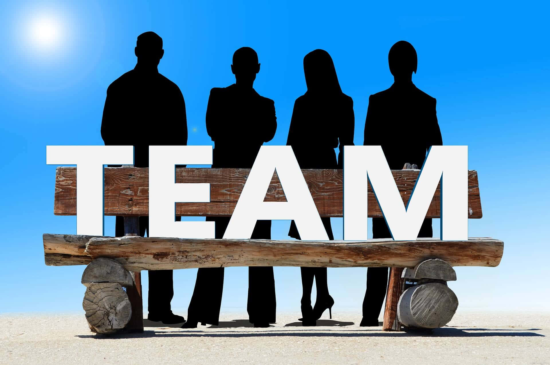 Teamwork 桌布 壁紙 — 四個商業剪影站在一張木凳後面，凳上有粗體白色字母拼成的TEAM字樣，在明亮的陽光下。