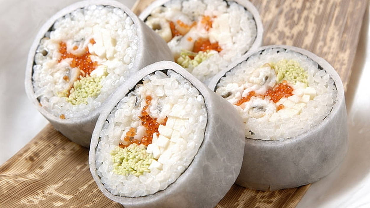 烹飪 桌布 壁紙 — 四個完美切好的壽司卷，配有米飯、飛魚籽、酪梨和白身魚，放在竹蓆上。