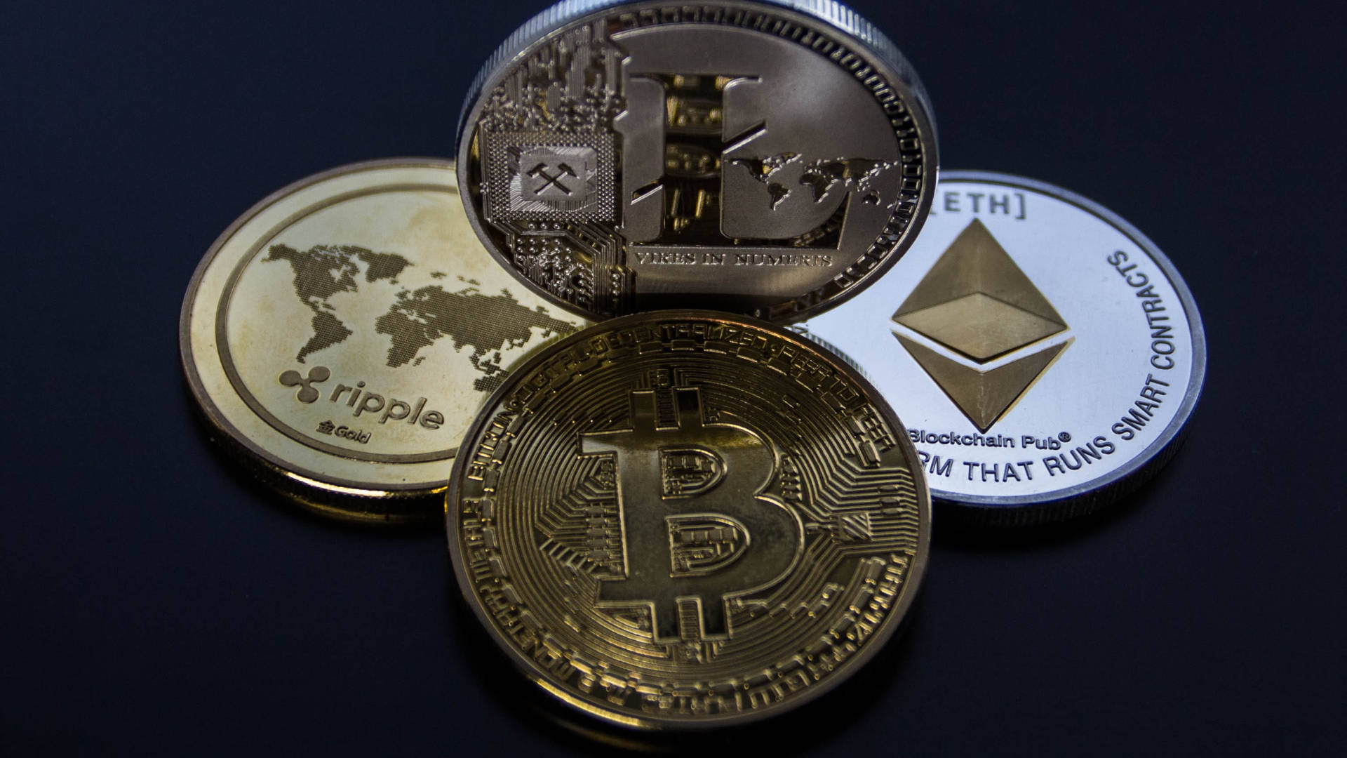 以太坊 桌布 壁紙 — 四個實體加密貨幣硬幣 Bitcoin Ethereum Ripple 和 Litecoin 排列在帶有金屬光澤和雕刻標誌的深色表面上。