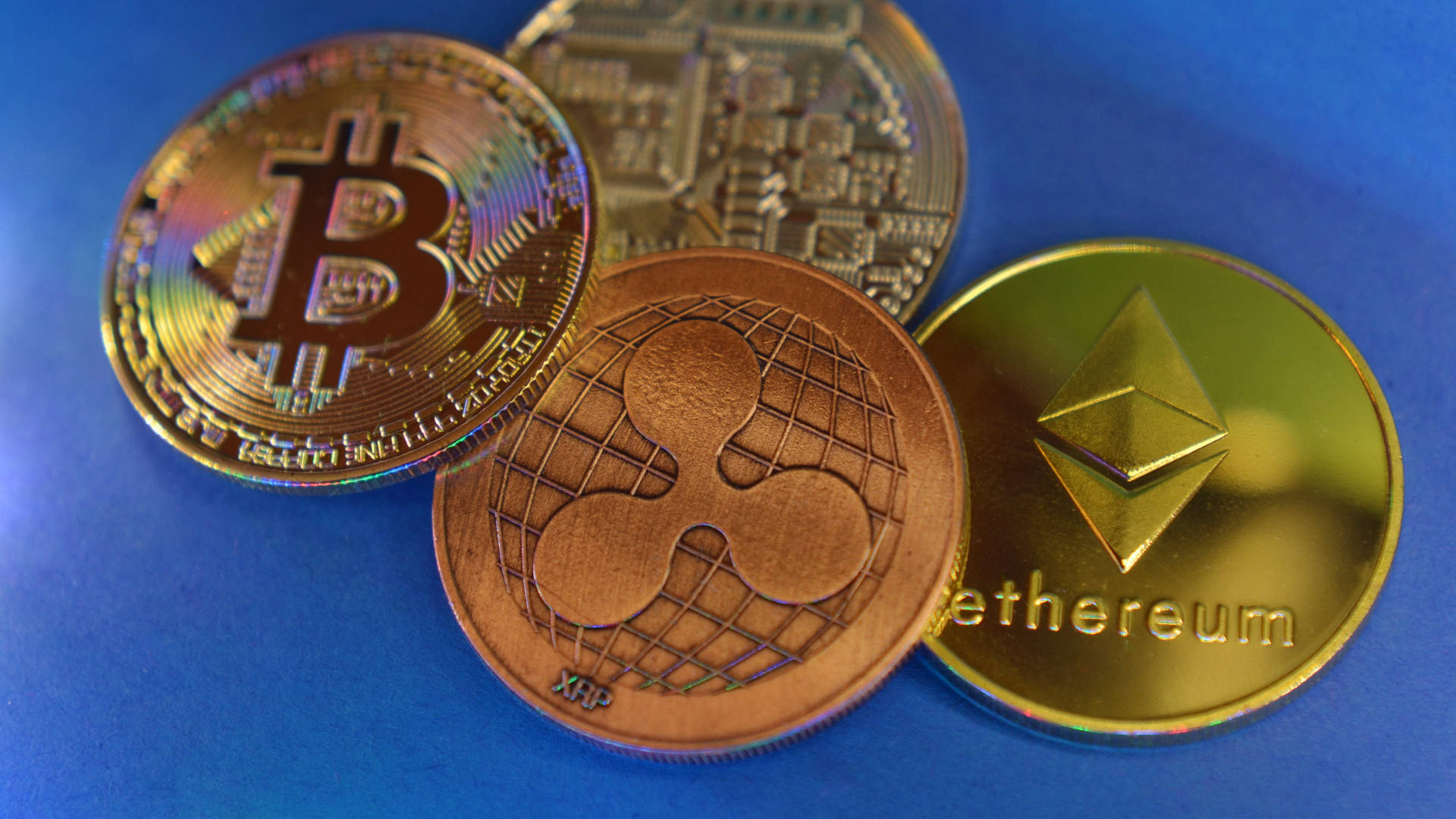 以太坊 桌布 壁紙 — 四個實體加密貨幣硬幣 Bitcoin Ethereum XRP 和 Litecoin 在明亮燈光下放置在充滿活力的藍色表面上。