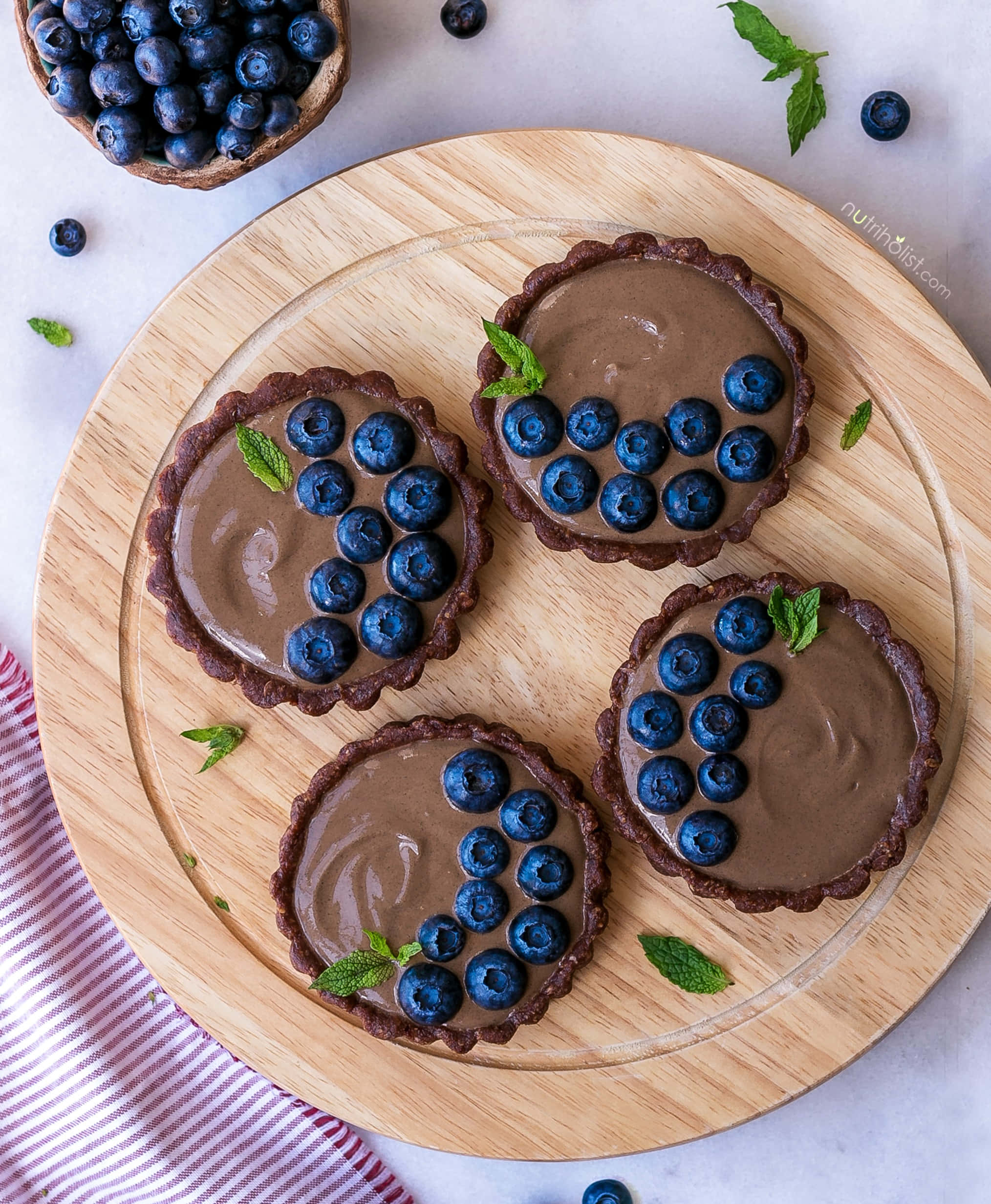 Blueberries Tart 桌布 壁紙 — 四個巧克力塔，上面放上新鮮藍莓和薄荷葉，放在一個木托盤上，旁邊有一塊條紋布。