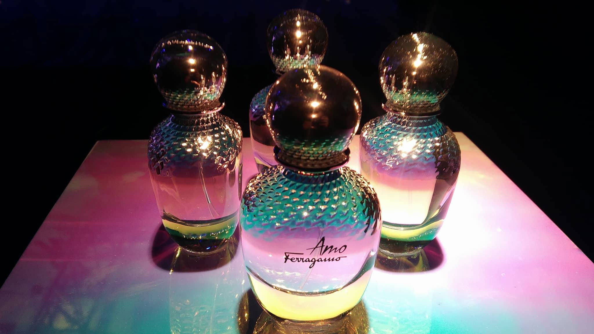Ferragamo 桌布 壁紙 — 四個帶有紋理玻璃和金屬蓋的彩虹色香水瓶，在反光表面上於彩色背光下閃耀。