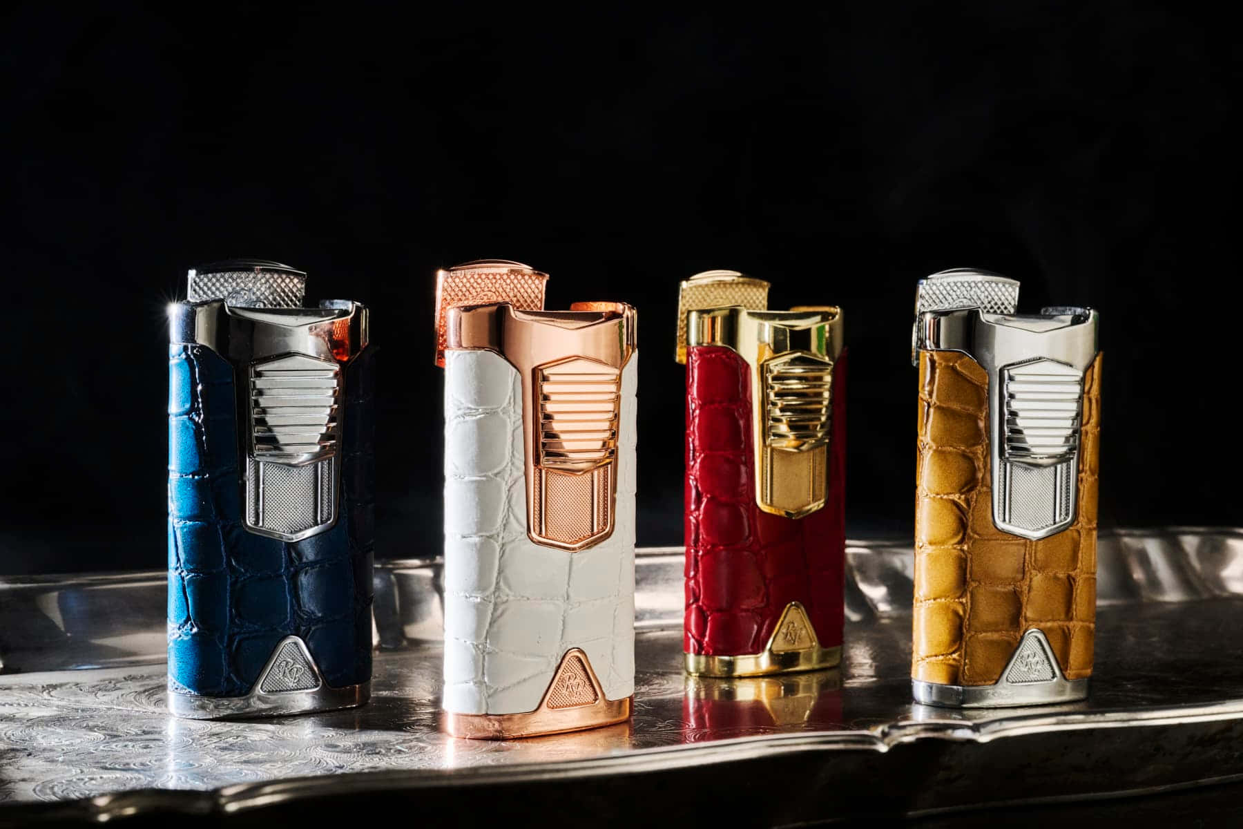 lighter 桌布 壁紙 — 四個帶有鱷魚皮和金屬裝飾的奢華雪茄打火機優雅地放置在黑色背景下的反光銀托盤上。