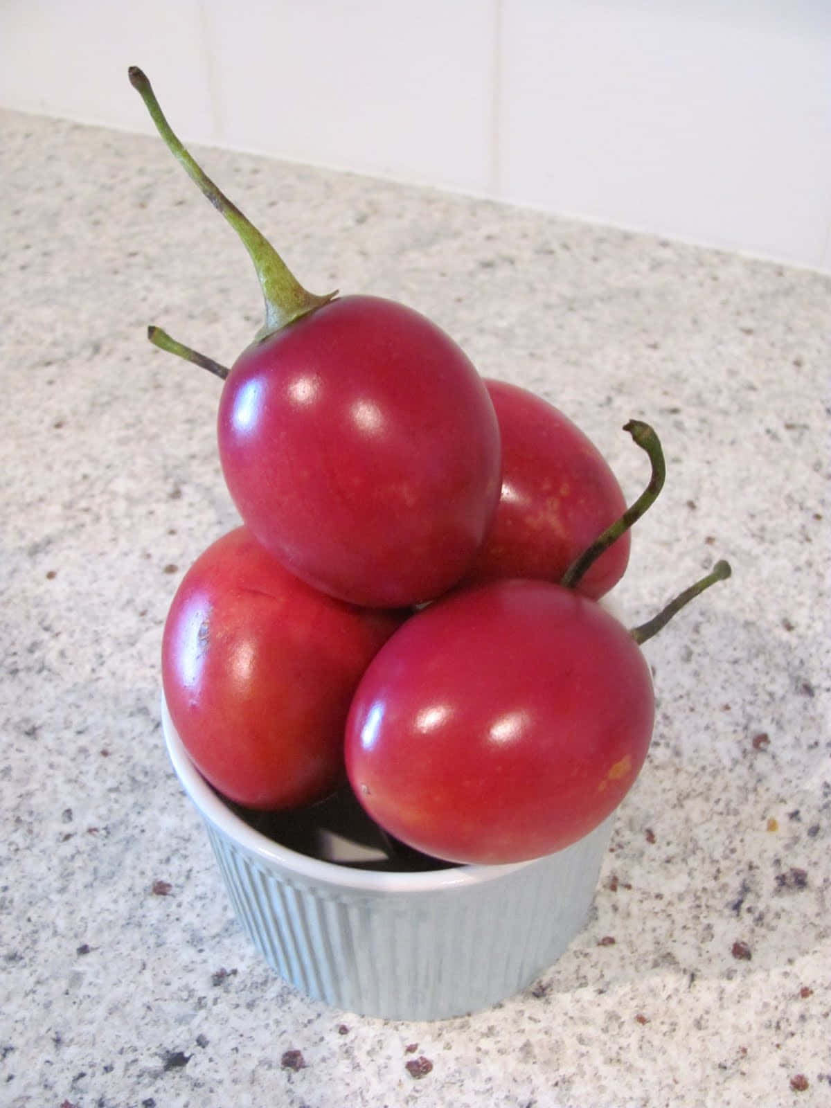 Fruit 桌布 壁紙 — 四個帶綠色莖的光澤紅番茄堆疊在一個白色帶槽紋的小烤碗中，放在有斑點的檯面上。