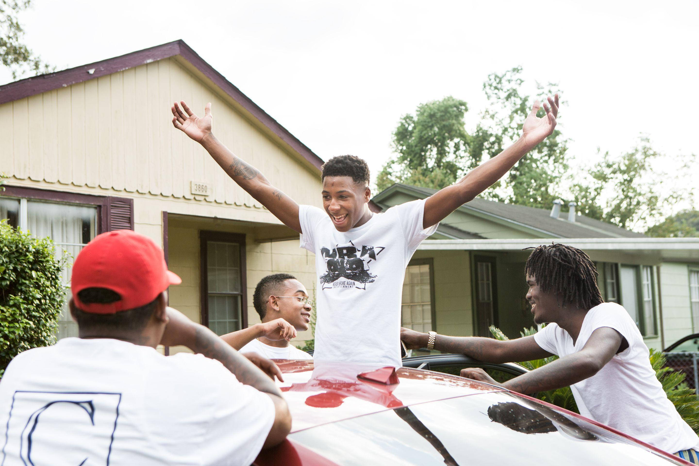 Youngboy 桌布 壁紙 — 四個年輕男子在一個有房屋和樹木的郊區社區中，開心地在一輛紅色汽車的引擎蓋上慶祝。