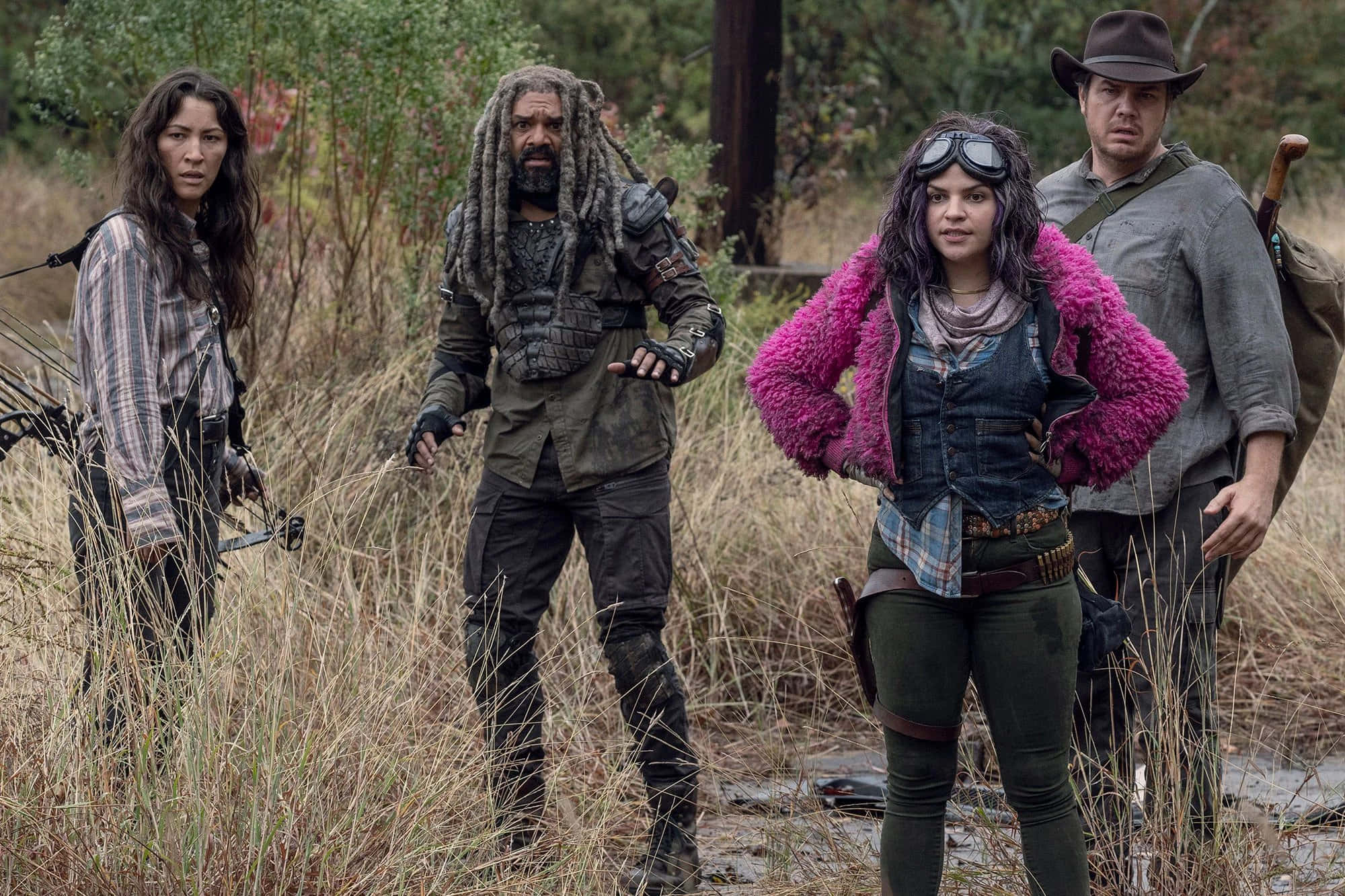 walking dead 桌布 壁紙 — 四個強悍的倖存者穿著末日後裝備，在多雲天空下的高乾草田中保持警覺。