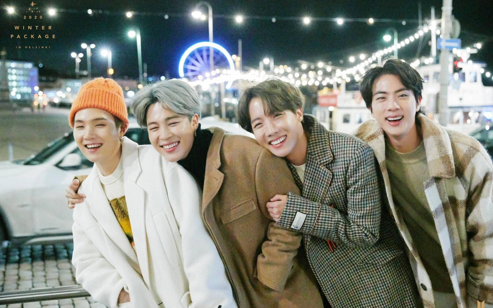 BTS 桌布 壁紙 — 四個微笑的年輕男子穿著舒適的冬季外套，在夜間於明亮的摩天輪和街燈附近一起擺姿勢。