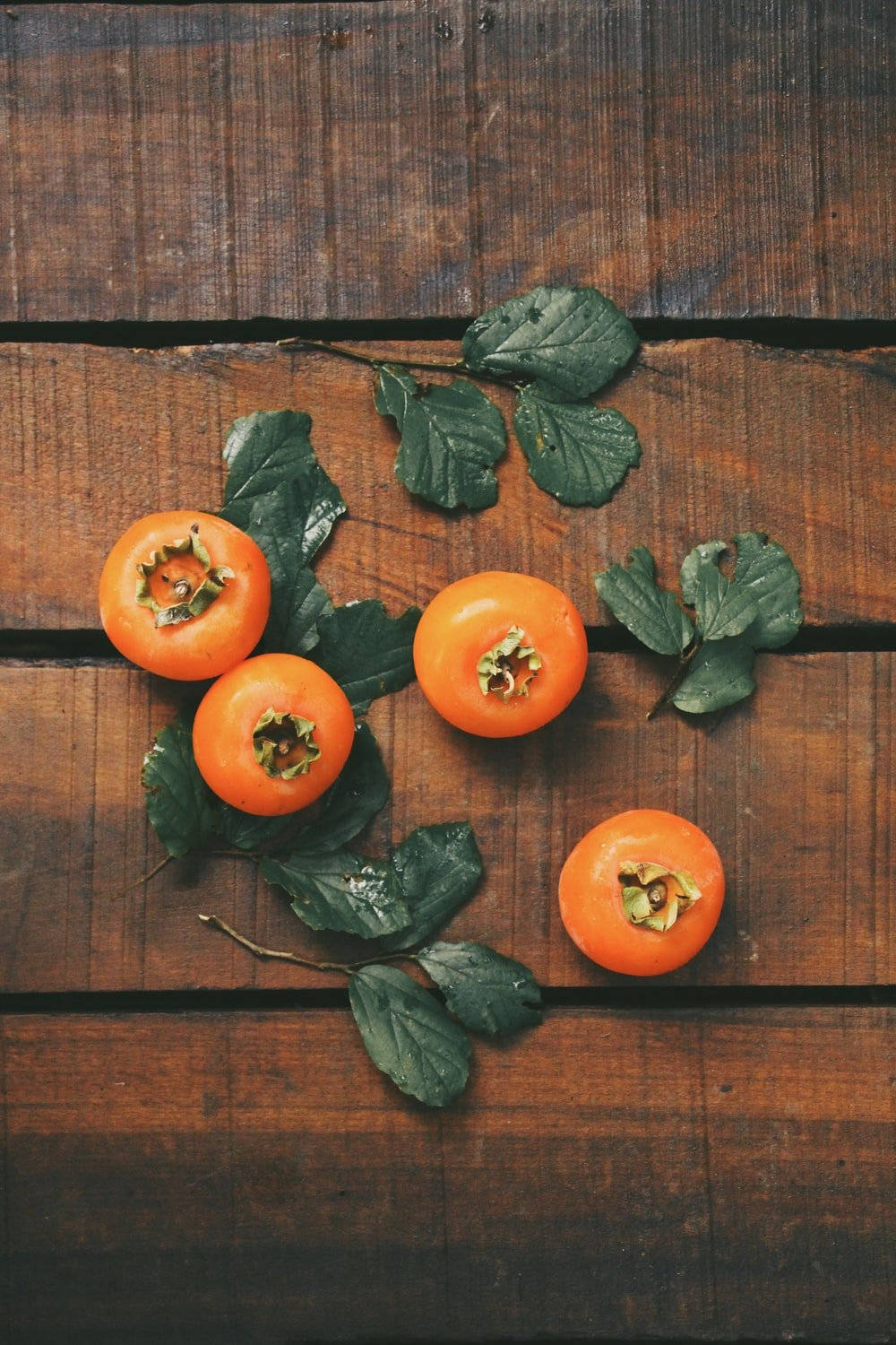 Fruit 桌布 壁紙 — 四個成熟的柿子帶著綠葉，放在經風化的木質表面上，呈現溫暖的秋色調。