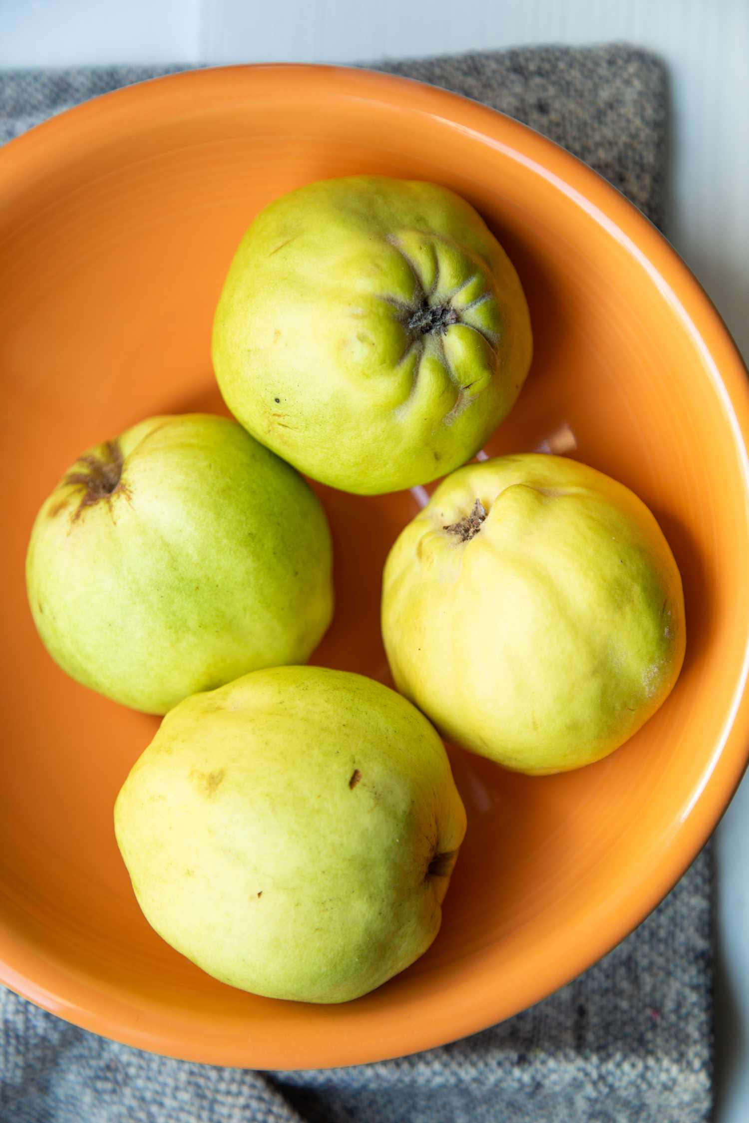 Quince 桌布 壁紙 — 四個成熟的榲桲放在一個鮮豔的橙色碗中，置於有紋理的灰色布上。