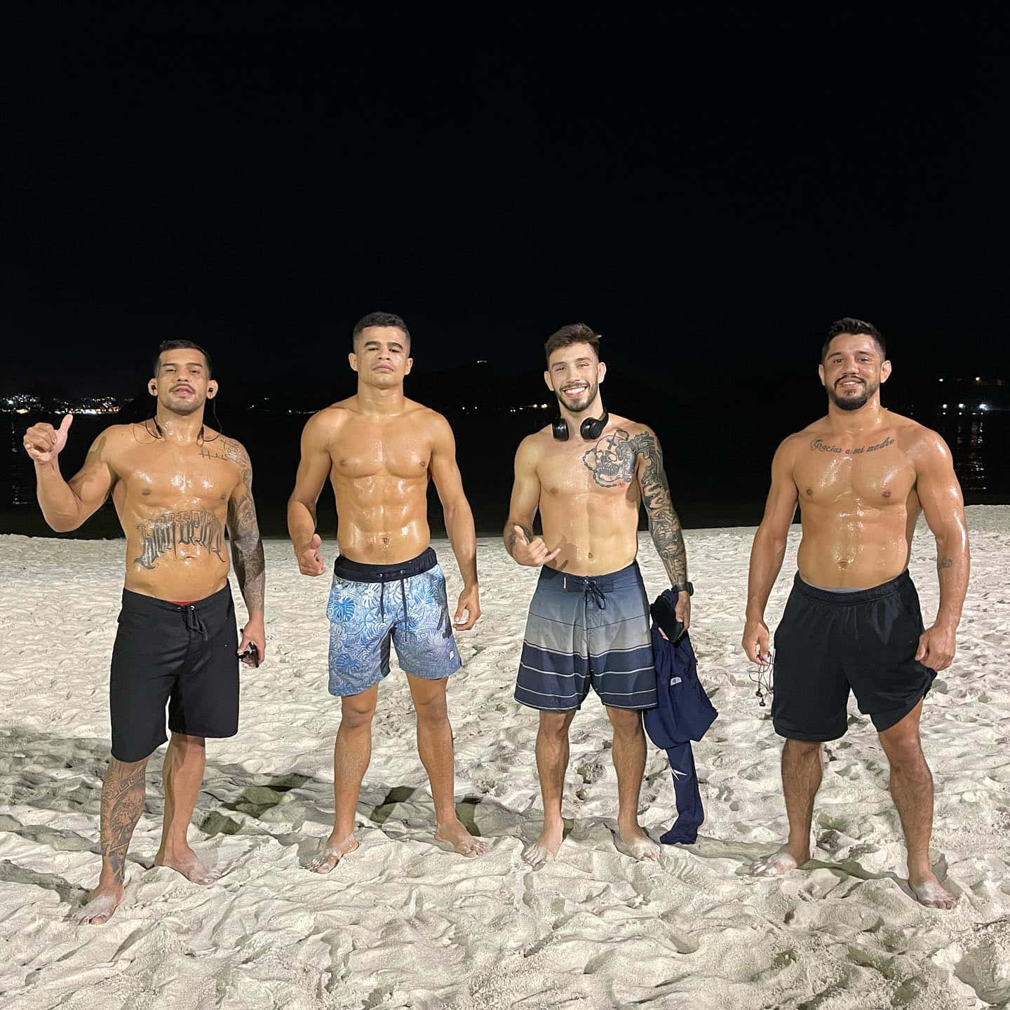 海灘 桌布 壁紙 — 四個有刺青的肌肉男在夜晚的城市燈光下赤腳站在沙灘上。