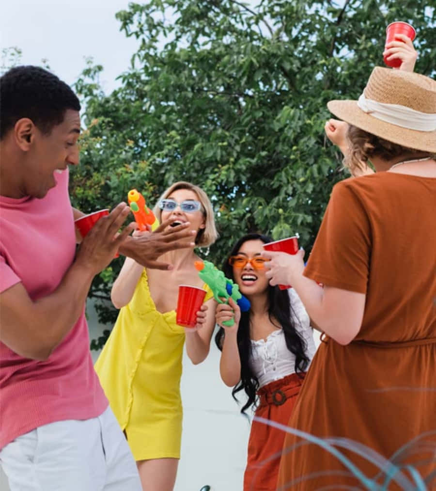 Party 桌布 壁紙 — 四個朋友在明亮的綠樹下笑著玩水槍捉人遊戲，手裡拿著紅色杯子。