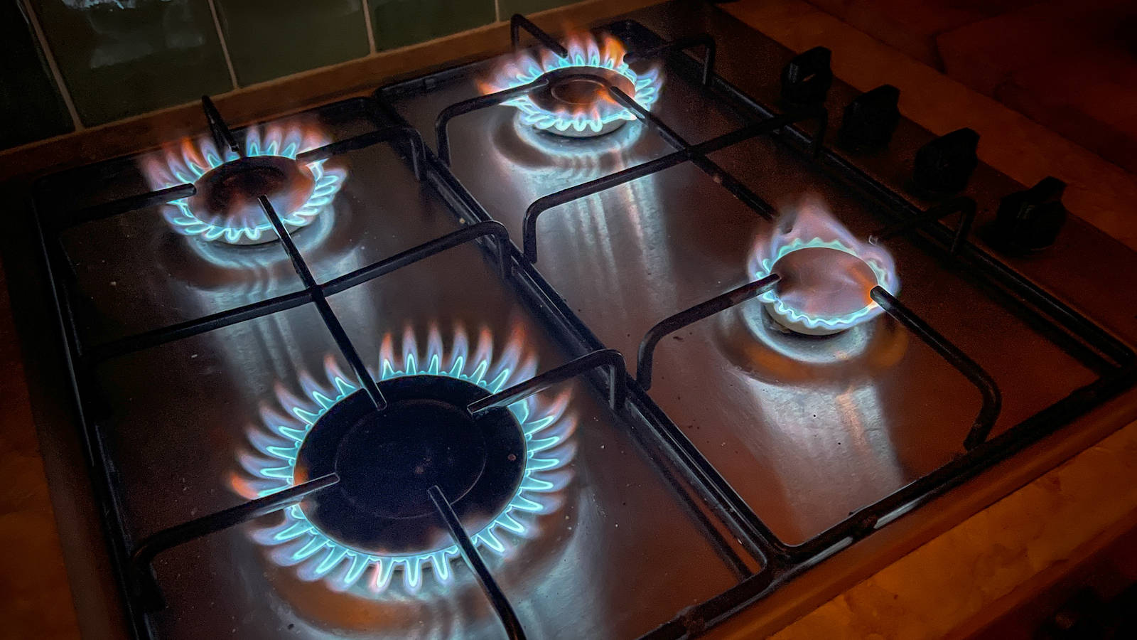 Gas 桌布 壁紙 — 四個瓦斯爐頭在不鏽鋼爐面上點燃，發出鮮豔的藍色火焰，廚房燈光昏暗。