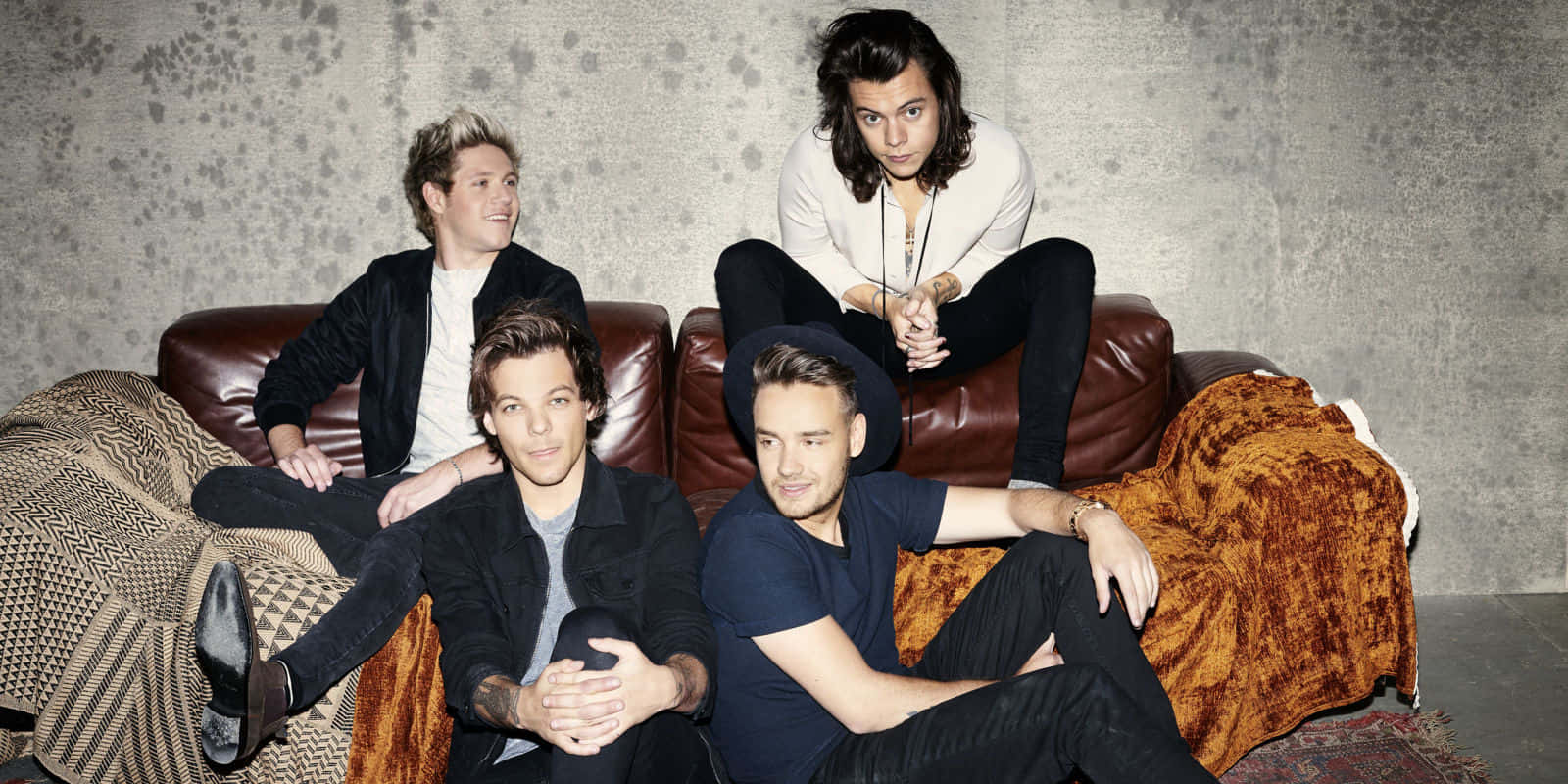 One Direction 桌布 壁紙 — 四個男人在有紋理的灰色牆前，隨意地躺在棕色皮沙發上，蓋著圖案毛毯。