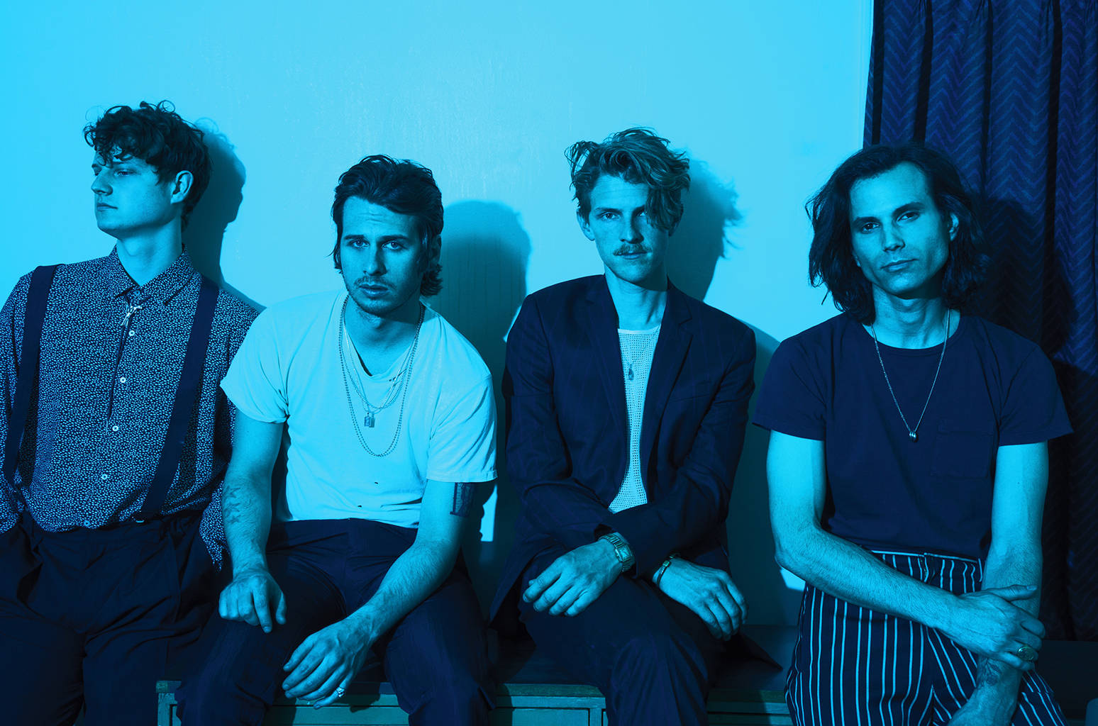 foster the people 桌布 壁紙 — 四個男人排成一排，坐在藍色牆壁前，在冷色燈光下，各有不同的風格和表情。