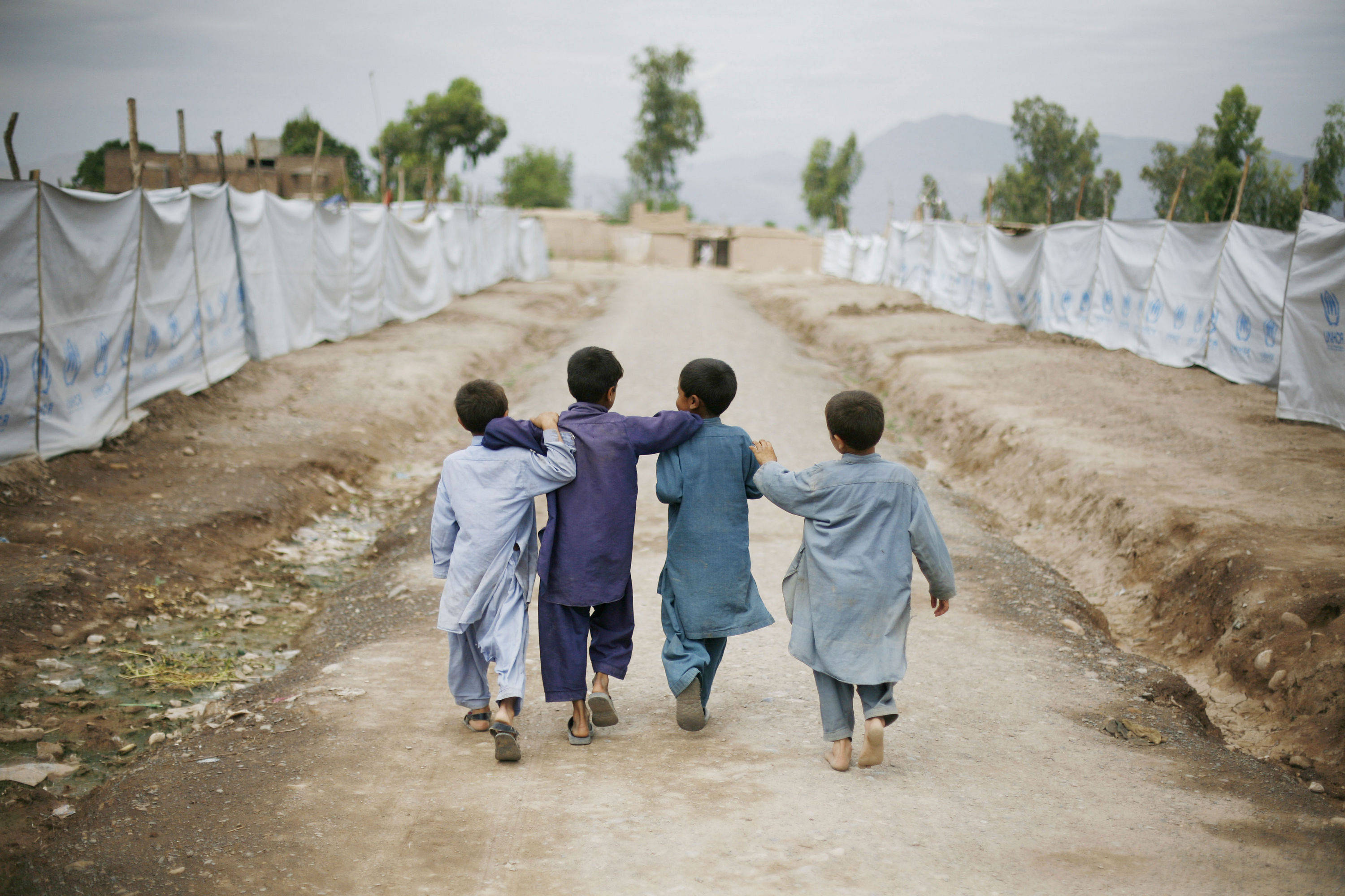 Afghanistan 桌布 壁紙 — 四個男孩臂挽著臂，走在一條塵土飛揚的鄉間小路上，路旁掛滿白色聯合國兒童基金會橫幅，天空陰沉。