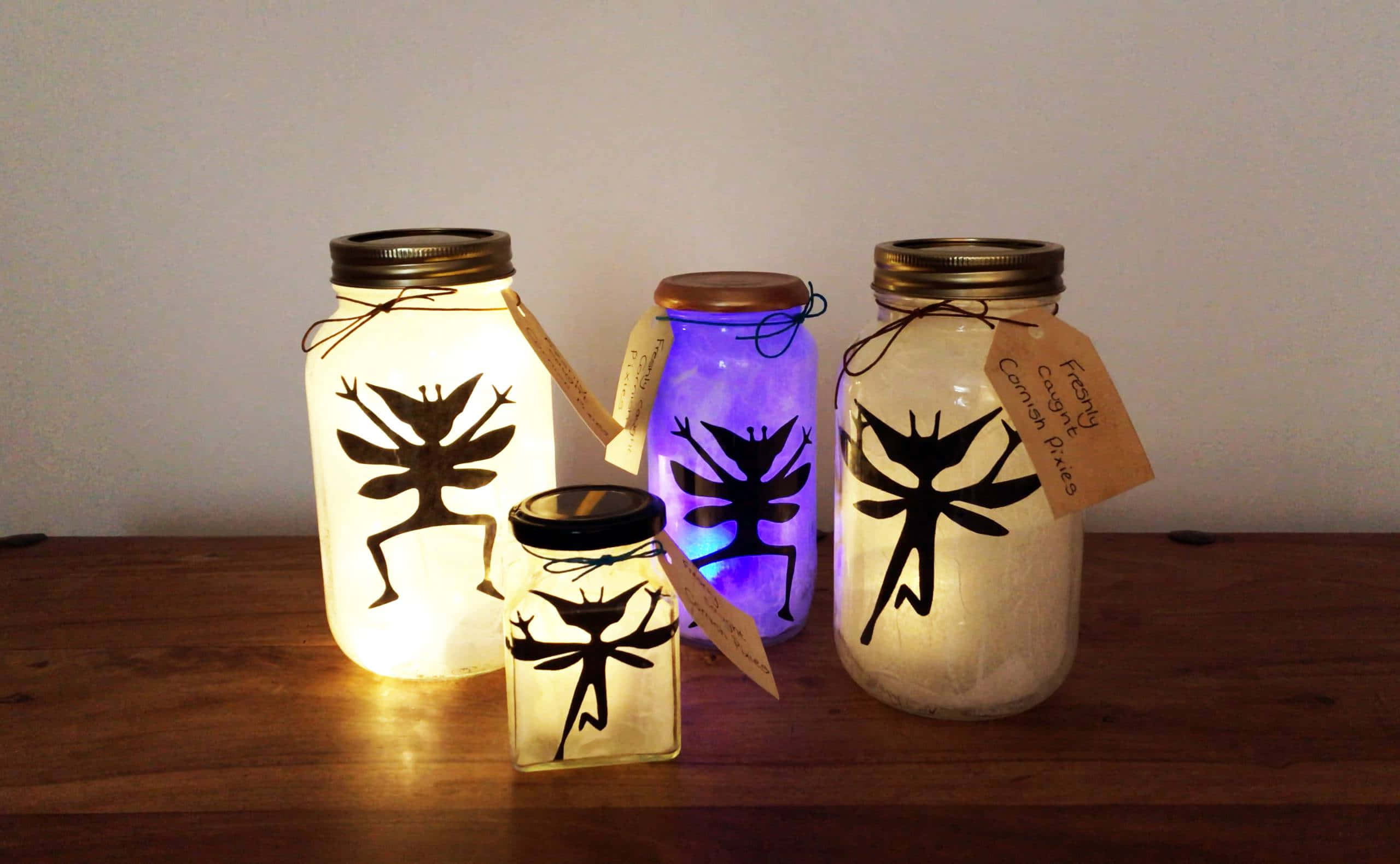 Light Up 桌布 壁紙 — 四個發光的玻璃瓶帶有仙女剪影，在素牆前將溫暖的光線投射在木質表面上。