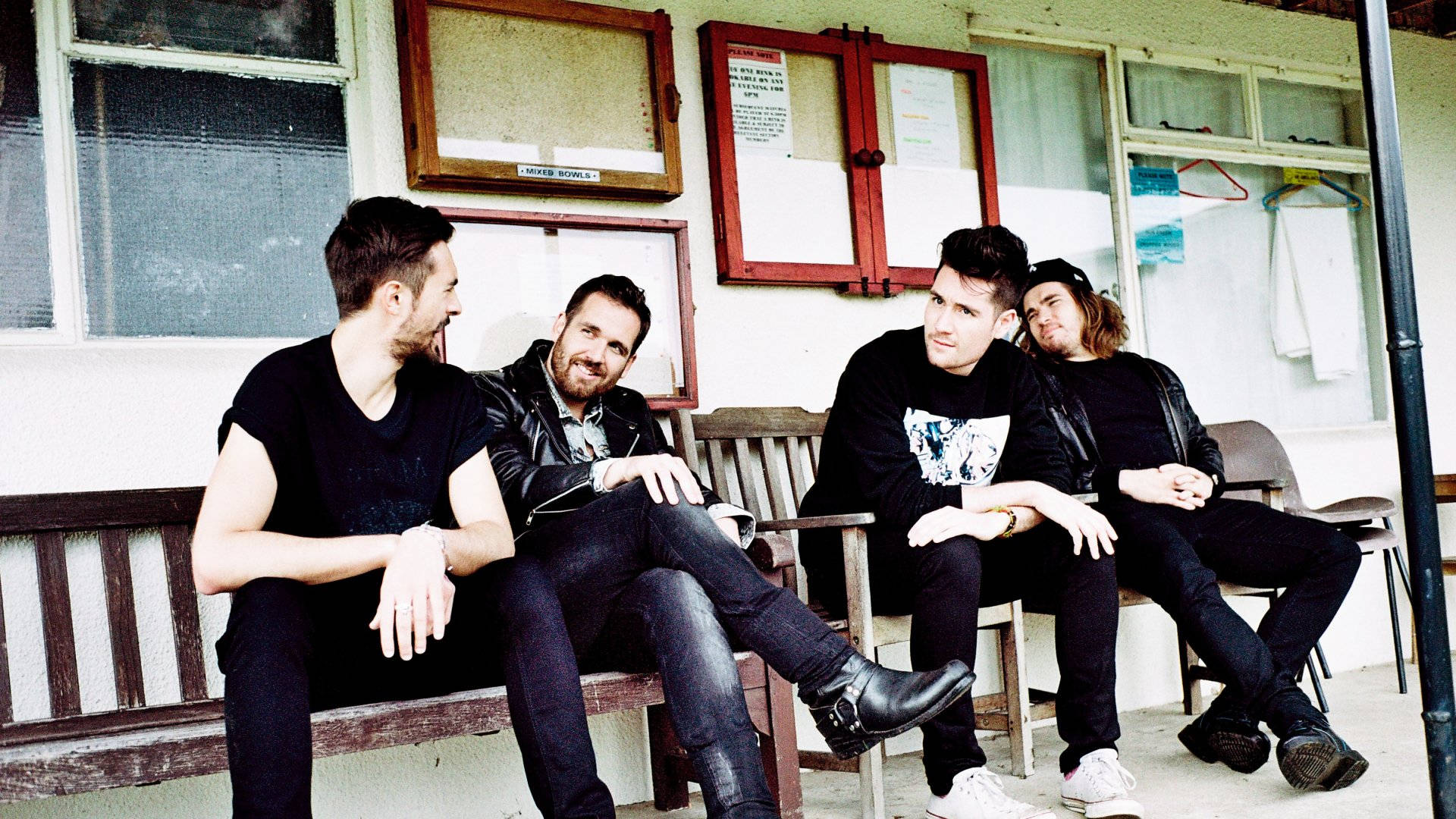 Bastille 桌布 壁紙 — 四個穿著休閒的男人放鬆地坐在一棟有紅色窗框的風化建築外的木長椅上。