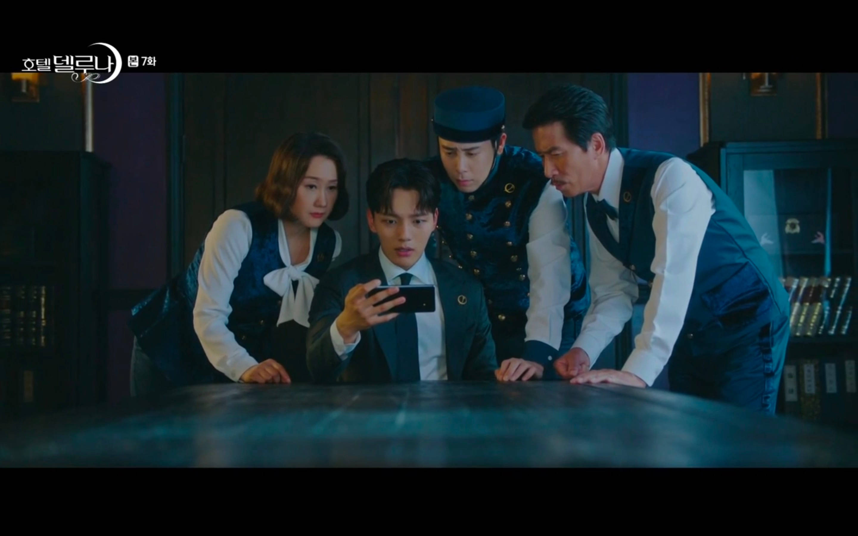 drama 桌布 壁紙 — 四個穿著復古制服的人俯身在一張桌子上，專注地盯著一部電話，房間燈光昏暗。