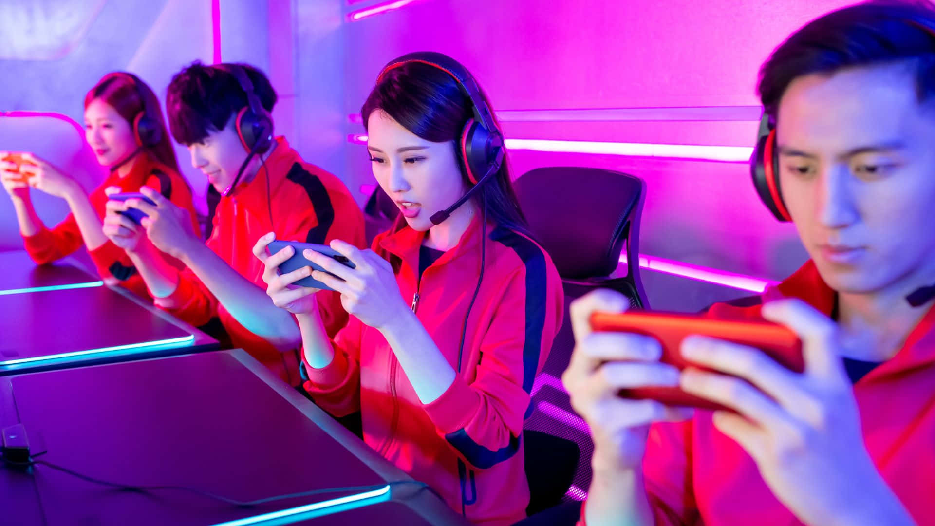 Gaming Tournaments 桌布 壁紙 — 四個穿著紅色外套的年輕玩家在充滿活力的粉紅和藍色霓虹燈下專注於他們的智慧型手機。