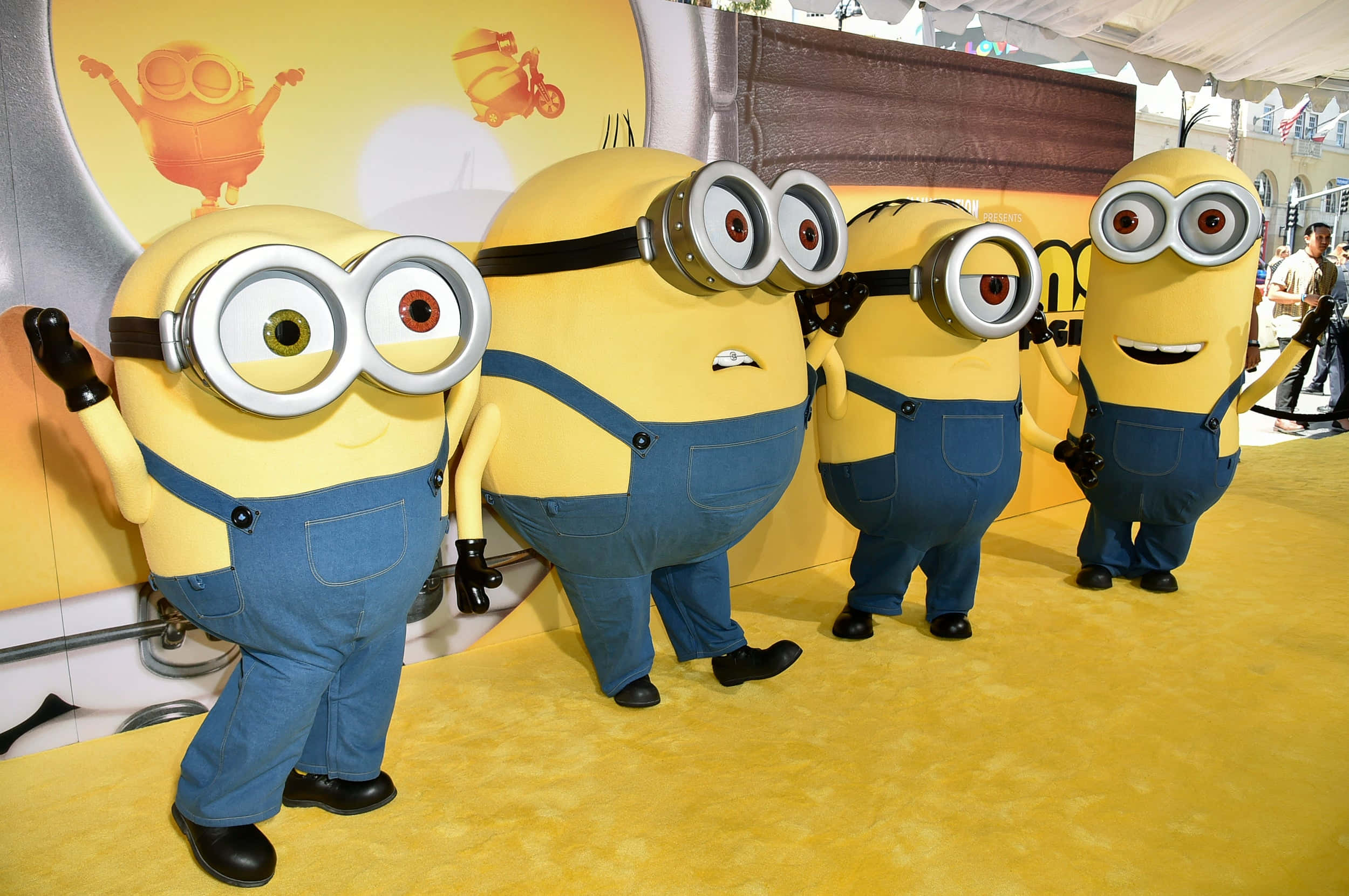 Minion 桌布 壁紙 — 四個穿著藍色工裝褲和護目鏡的黃色小小兵在促銷活動中開心地擺姿勢在亮黃色地毯上。