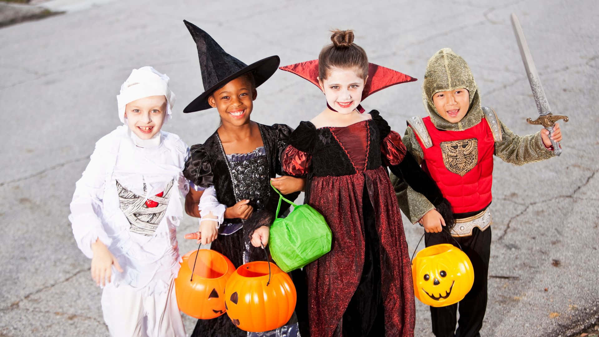 Halloween Costumes 桌布 壁紙 — 四個笑臉孩子在萬聖節服裝中拿著南瓜桶站在鋪砌的街道上。