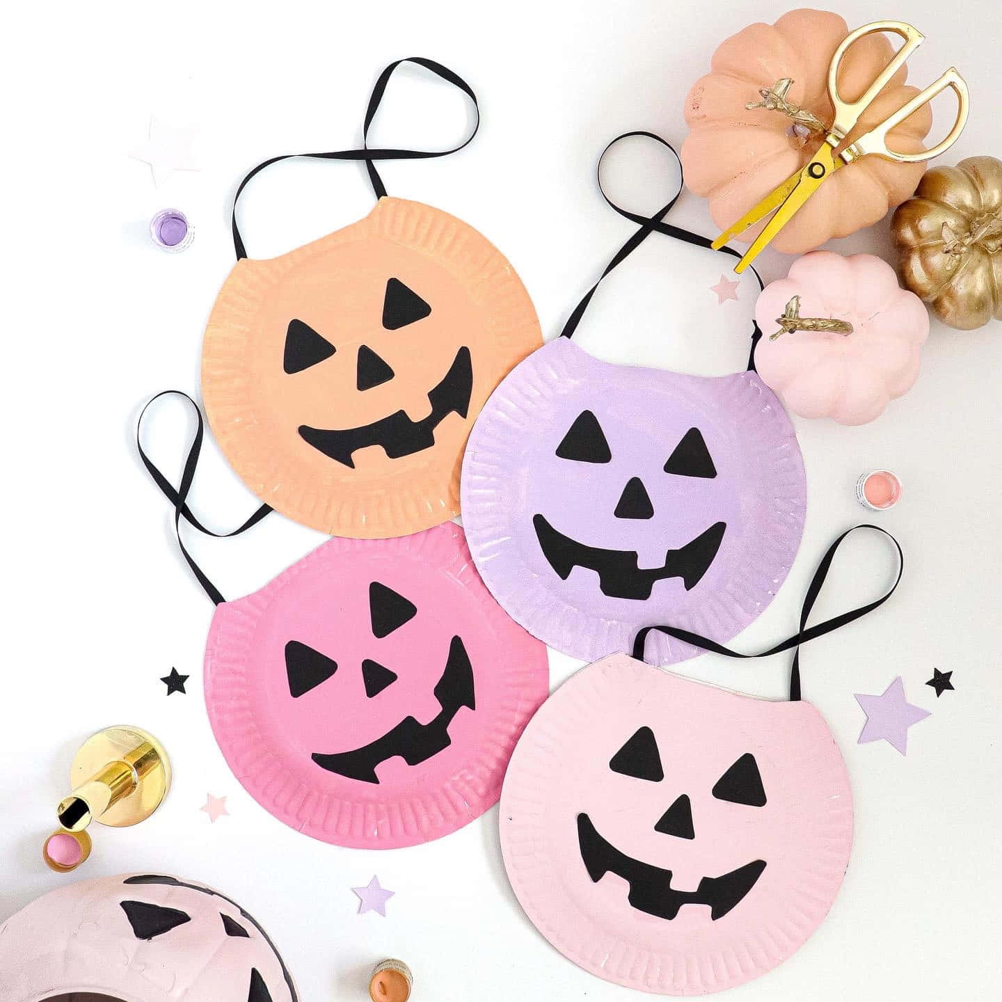 Halloween Crafts 桌布 壁紙 — 四個粉彩色紙盤南瓜燈面具，帶有黑色三角形眼睛和笑容，排列在白色表面上，配有裝飾性南瓜和剪刀。