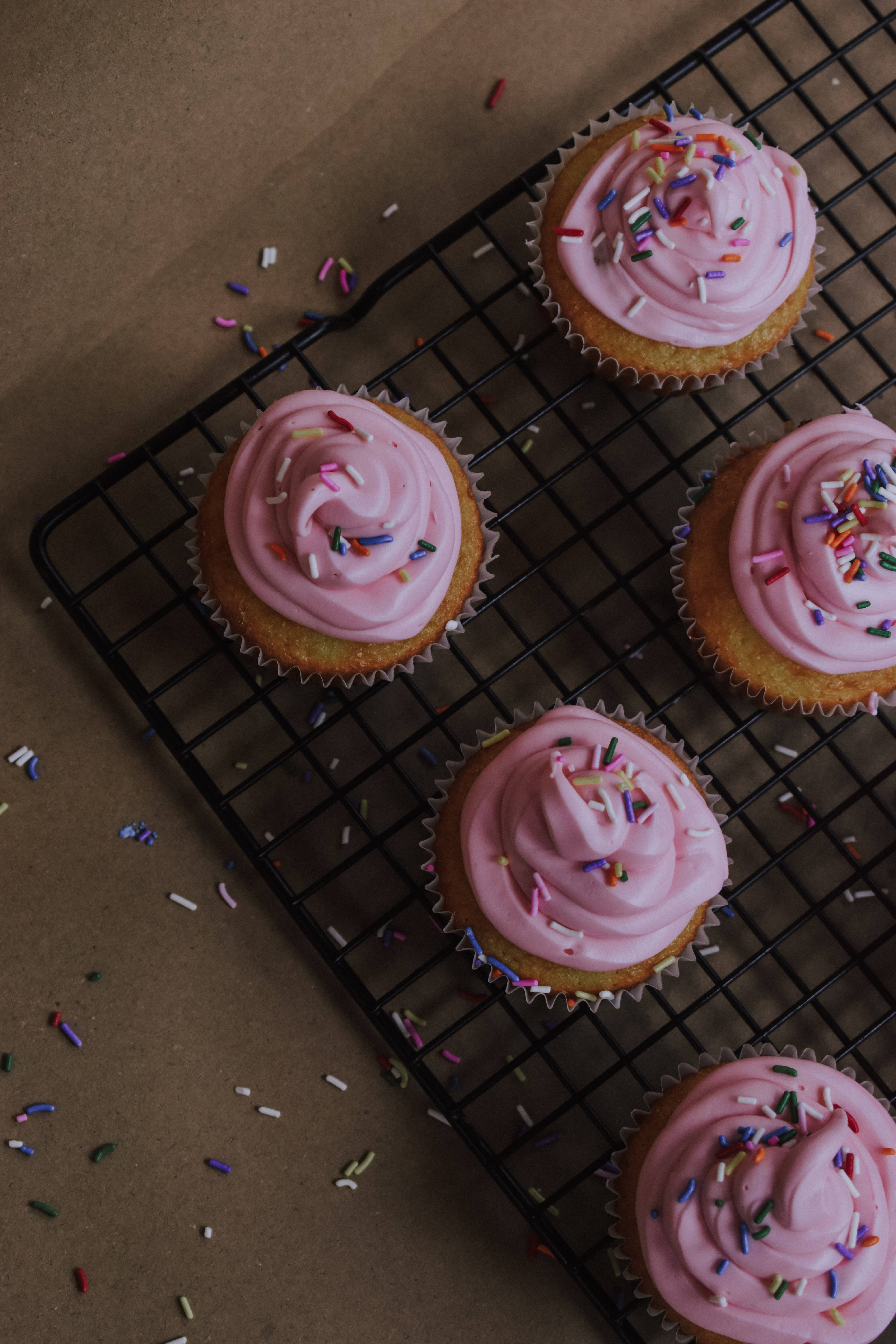 Cupcake 桌布 壁紙 — 四個粉紅色糖霜配彩色糖粒的杯子蛋糕放在棕色紙上的黑色金屬架上。