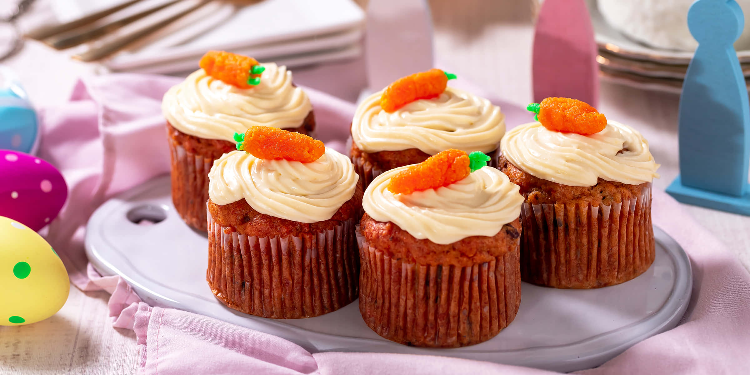 Cupcake 桌布 壁紙 — 四個胡蘿蔔蛋糕杯子蛋糕，上面有奶油乳酪糖霜和小胡蘿蔔裝飾，放在一個盤子上，旁邊有彩色復活節彩蛋。