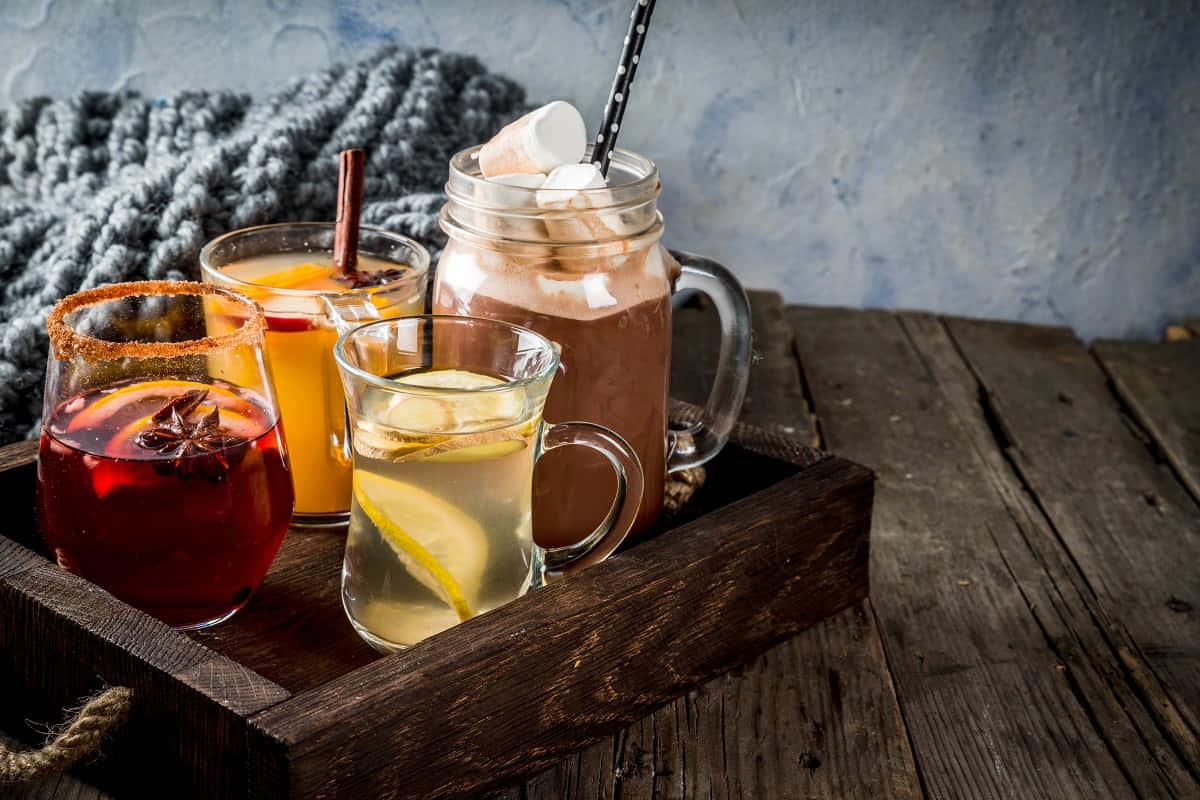 Winter Drinks 桌布 壁紙 — 四個舒適的飲品放在梅森瓶和玻璃杯中，置於一個質樸的木托盤上，旁邊有一條編織毯子。