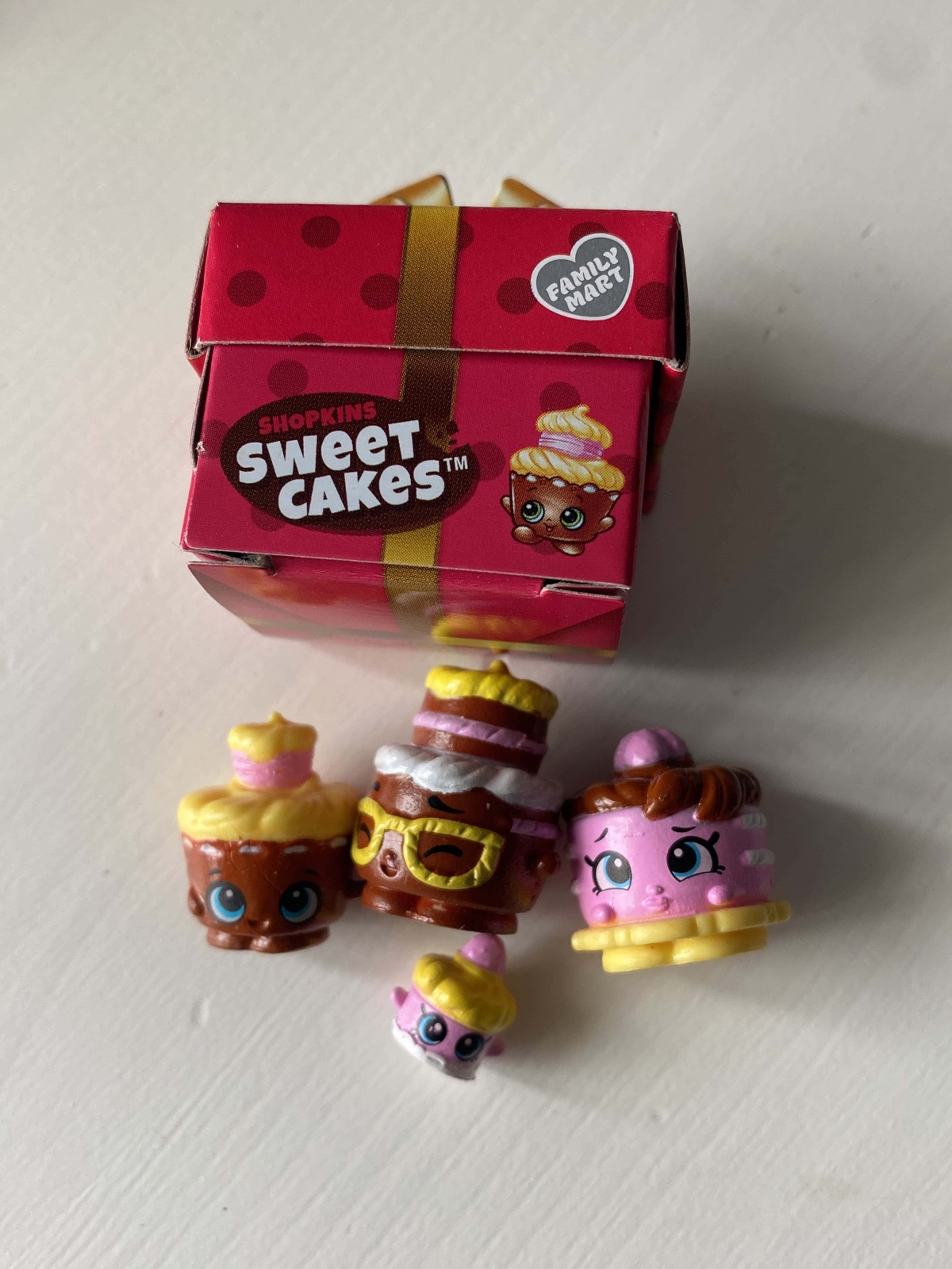 Shopkins 桌布 壁紙 — 四個色彩繽紛的Shopkins Sweet Cakes玩具放在白色表面上，旁邊是她們粉紅色圓點Family Mart盒子。
