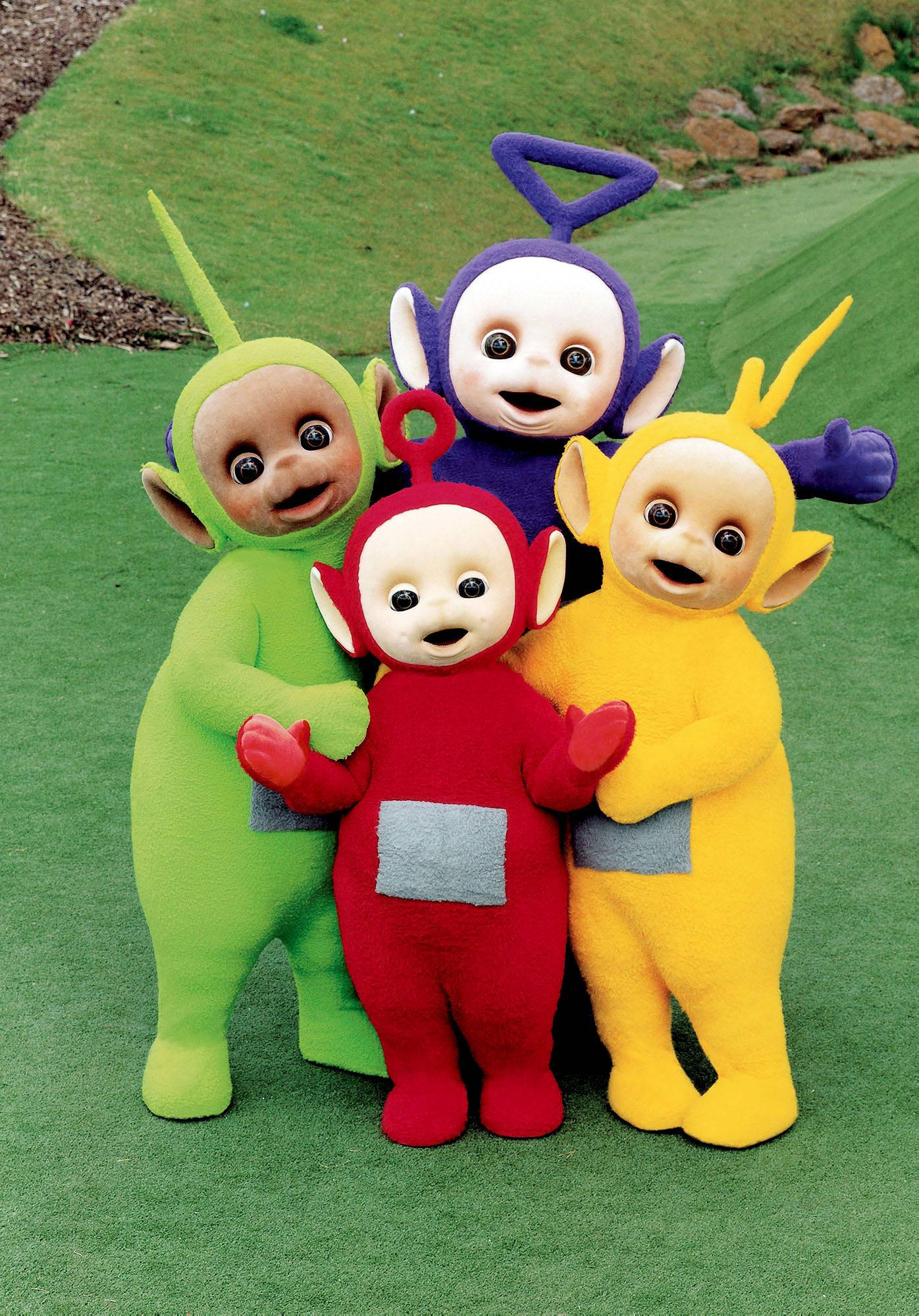 Cartoon 桌布 壁紙 — 四個色彩繽紛的Teletubbies穿著綠色、紅色、紫色和黃色的服裝，帶著喜悅的表情擠在一個草地上。