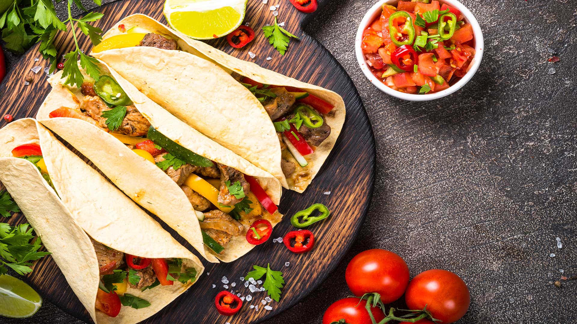 Mexican 桌布 壁紙 — 四個色彩繽紛的墨西哥捲餅，餡料有肉、甜椒和香草，放在深色木板上，旁邊有新鮮莎莎醬、番茄和萊姆角。