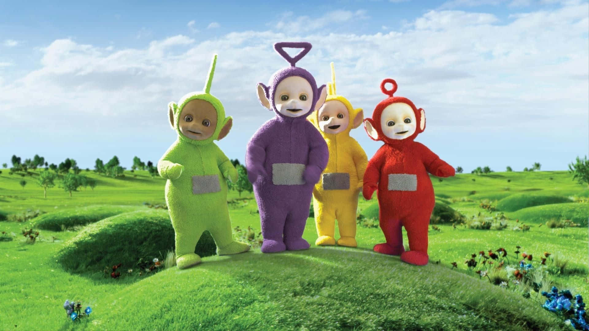 Teletubbies 桌布 壁紙 — 四個色彩繽紛的天線寶寶站在一個草地山坡上，頭頂是晴朗的藍天和 fluffy 雲。