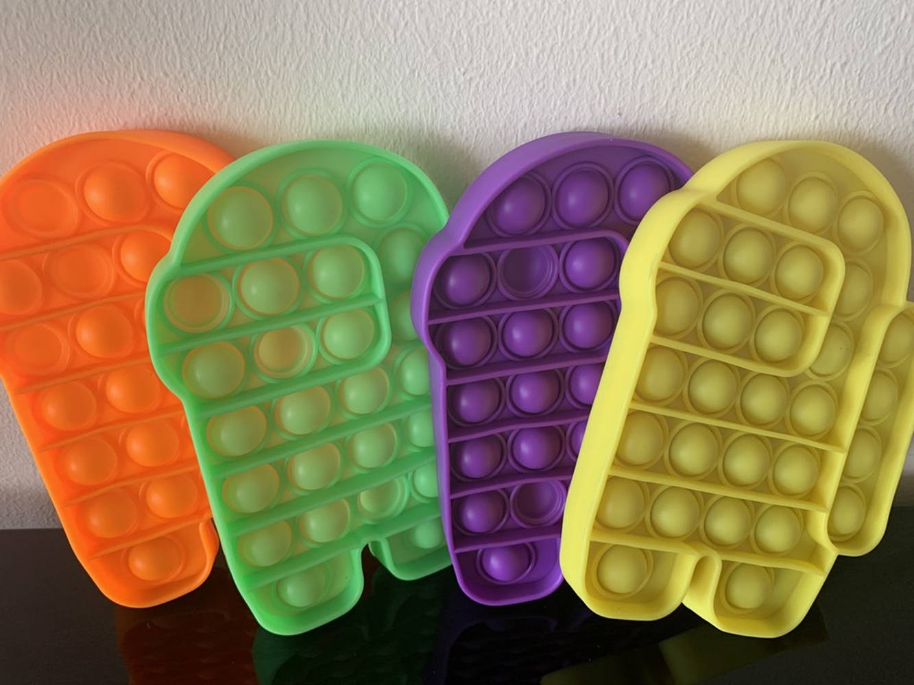 Fidget Toys 桌布 壁紙 — 四個色彩繽紛的彈出式減壓玩具，分別是橙色、綠色、紫色和黃色，靠在白色牆壁上，置於深色反光表面上。