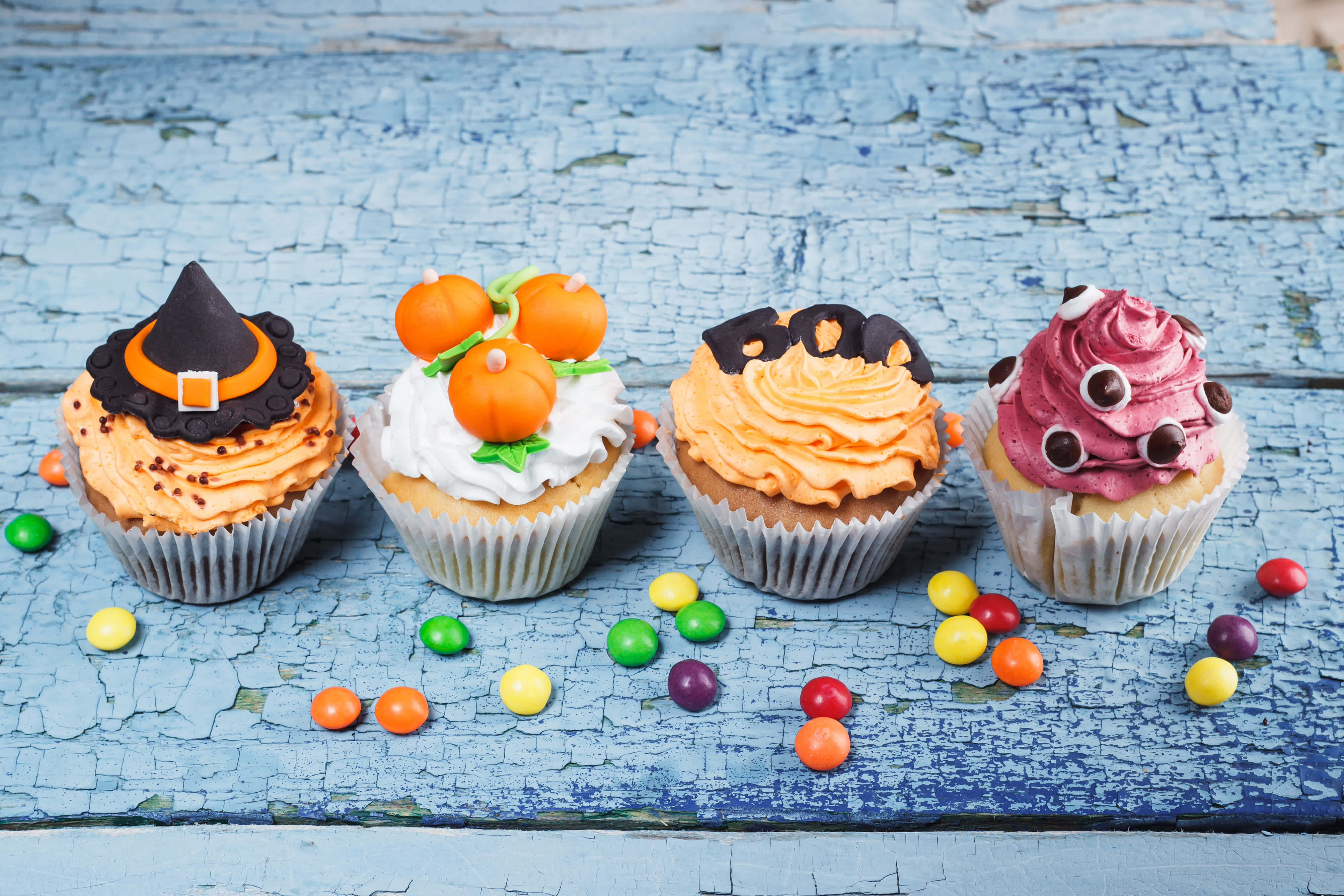 Halloween Cupcakes 桌布 壁紙 — 四個萬聖節主題杯子蛋糕，飾有巫婆帽、南瓜、鬼魂和怪獸眼睛，放在經風化的藍色木質表面上，周圍散落著色彩繽紛的糖果。