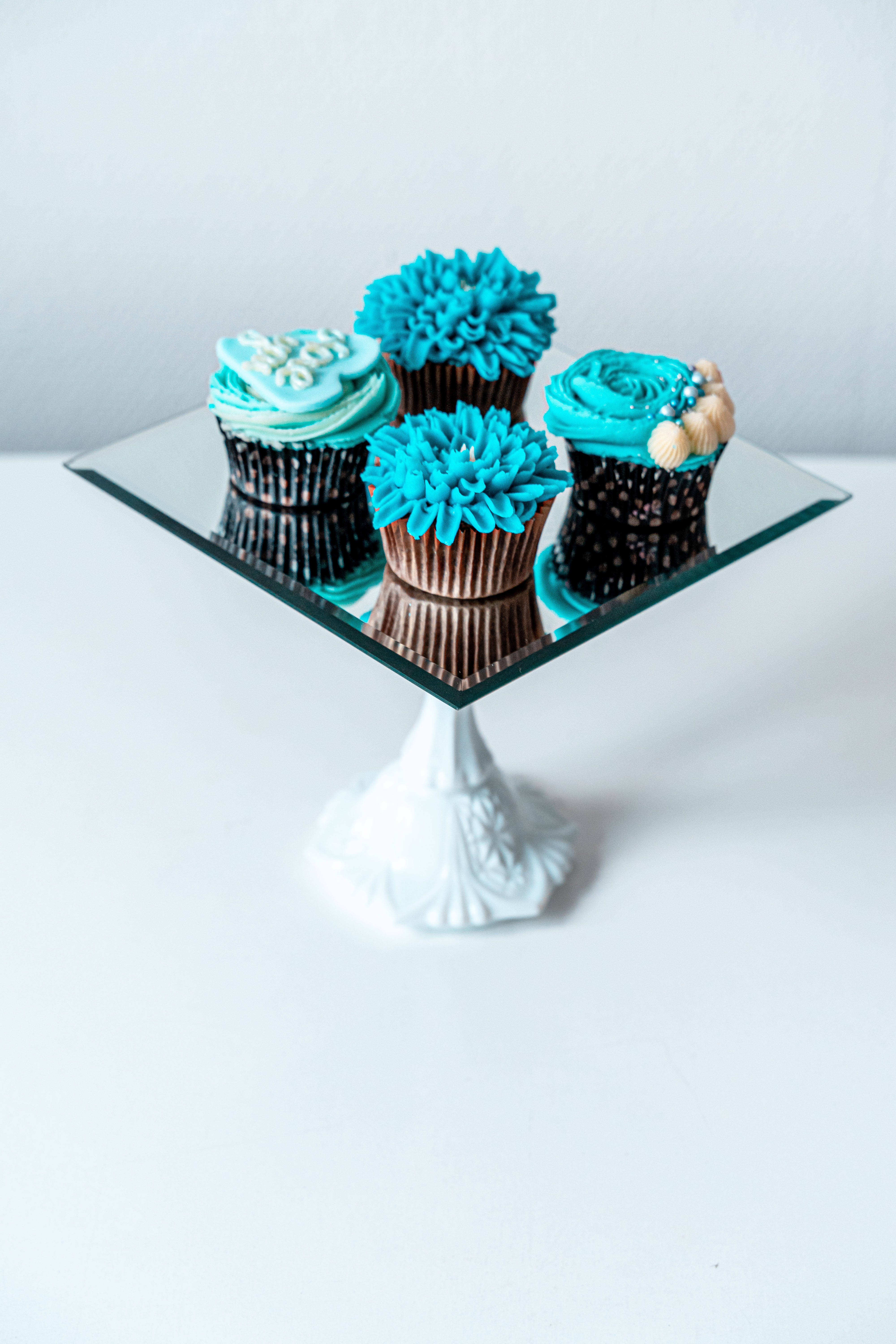 Cupcake 桌布 壁紙 — 四個藍綠色糖霜紙杯蛋糕，帶有裝飾性漩渦，優雅地放在一個帶白色華麗底座的鏡面玻璃蛋糕架上。