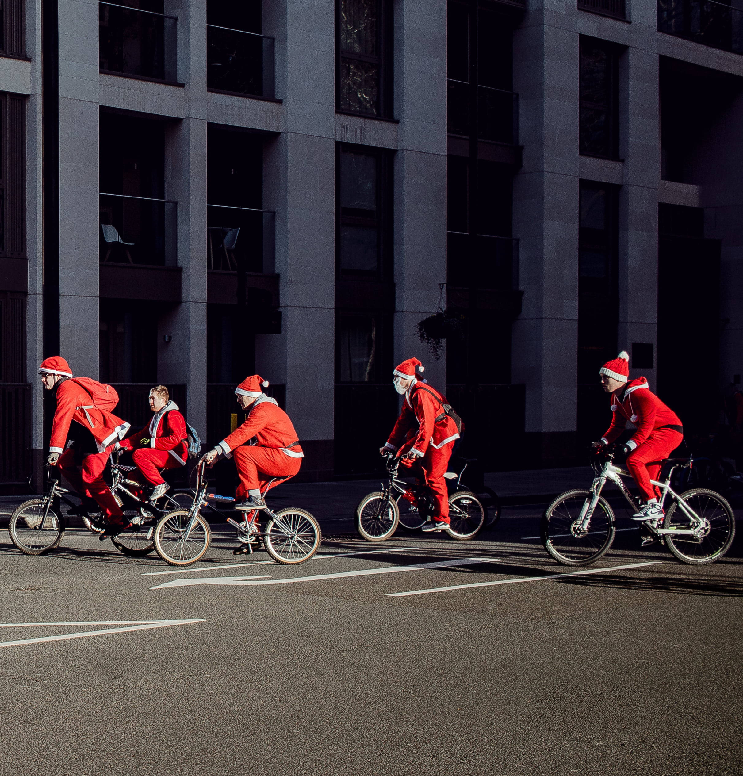 Santa Claus 桌布 壁紙 — 四個身穿全套紅色聖誕老人服裝的人騎著腳踏車，沿著一條現代城市街道行駛，背景是高聳的灰色建築物。