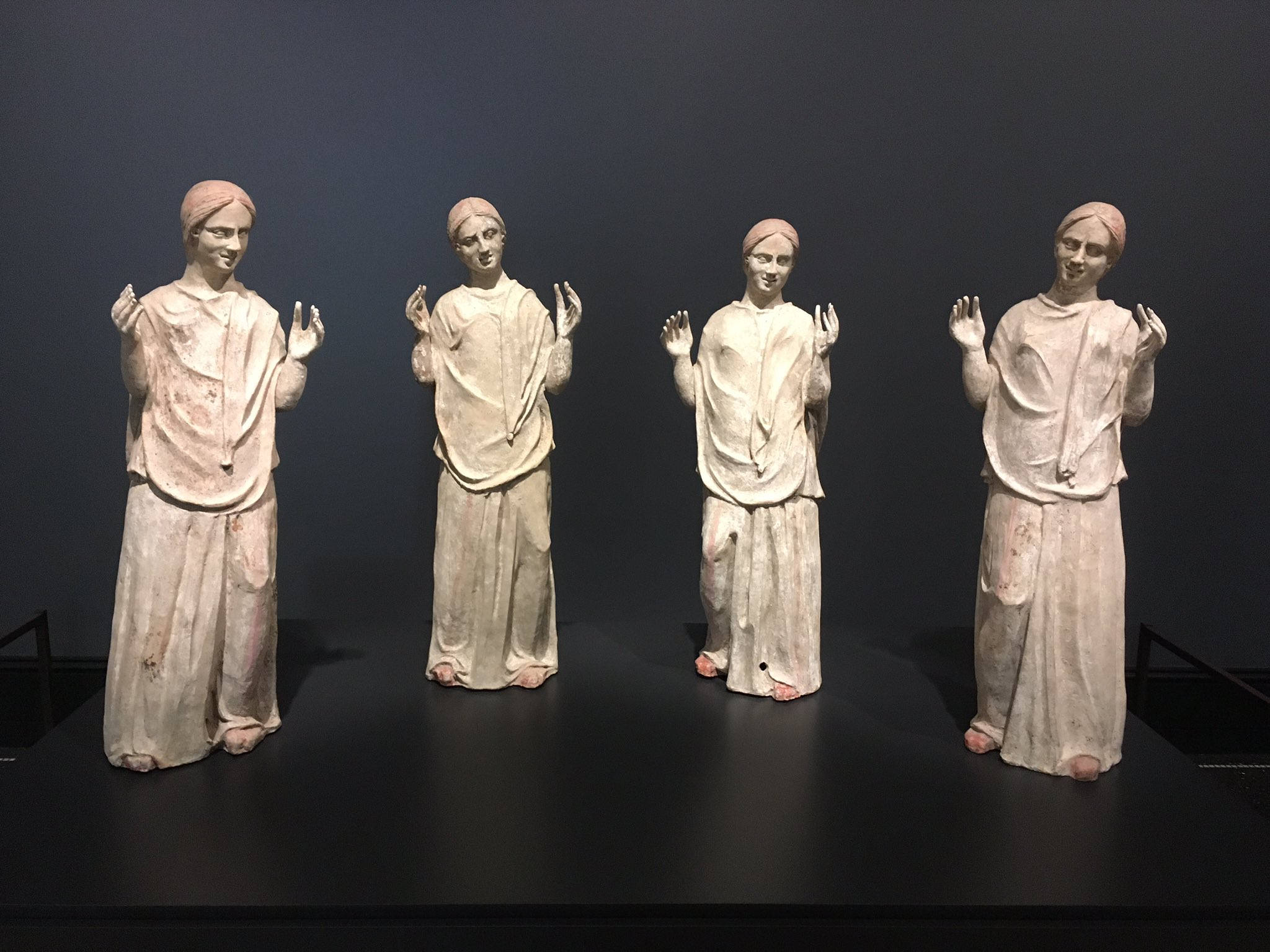 Museum 桌布 壁紙 — 四個身穿長袍、舉起雙手的相同古代雕像，在黑暗的畫廊牆前排成一排。
