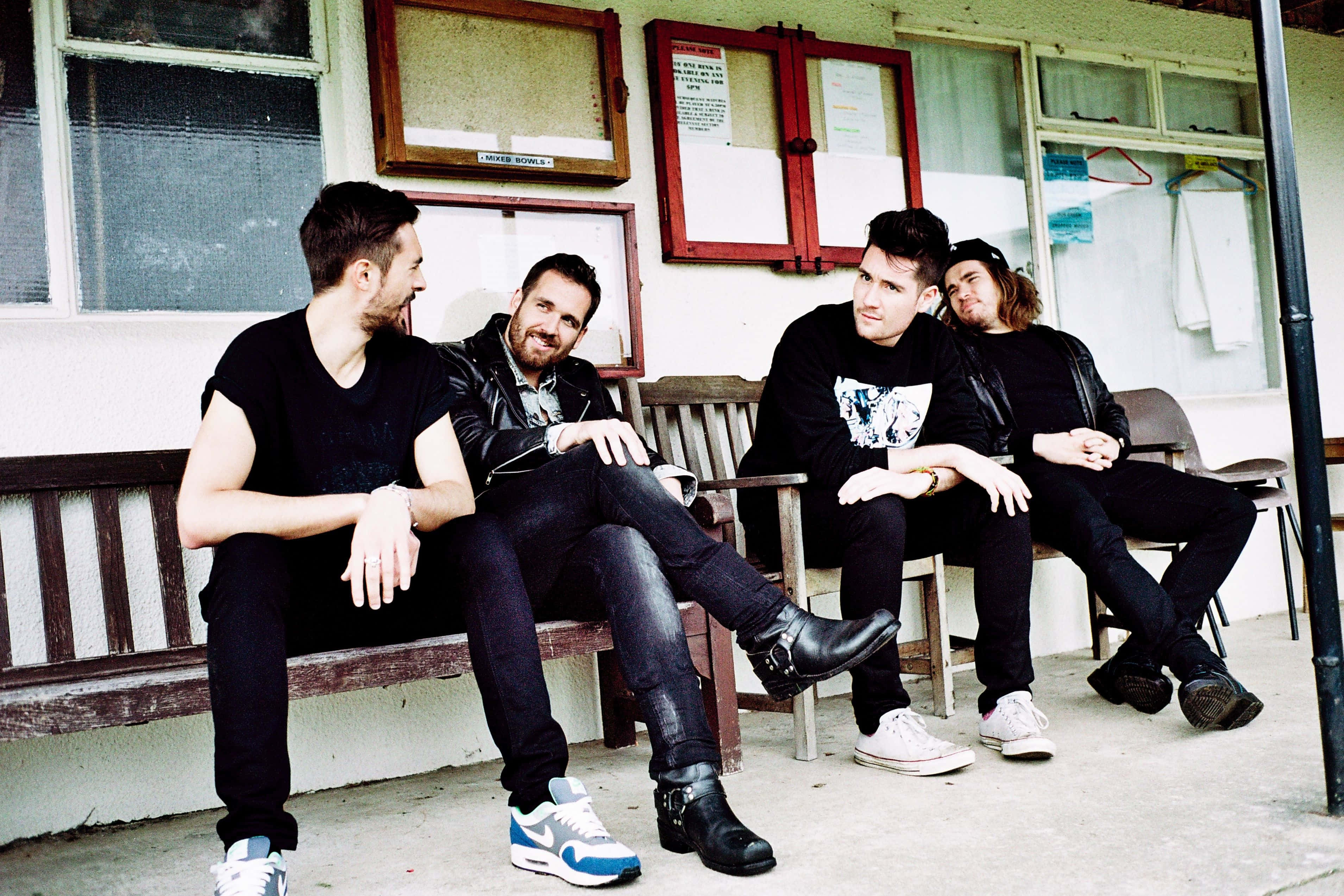 Bastille 桌布 壁紙 — 四個身穿黑色衣服的男人隨意地坐在一棟有紅色窗框的風化建築物外的木長椅上。