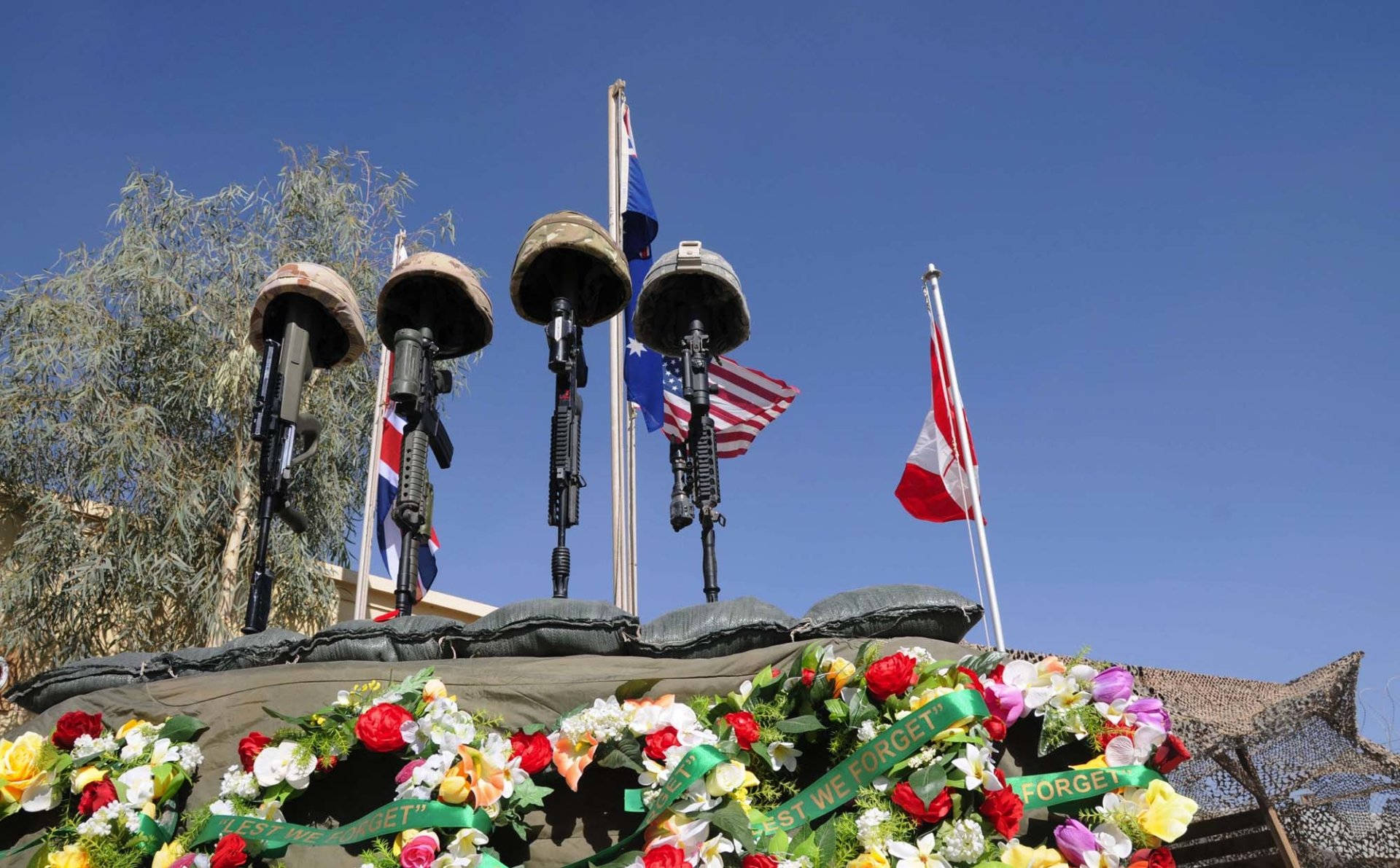 Veterans 桌布 壁紙 — 四個軍用頭盔安裝在步槍上，矗立在一個用沙袋堆成的紀念碑上，裝飾著色彩繽紛的花圈和飄揚的國旗，在晴朗的藍天下。