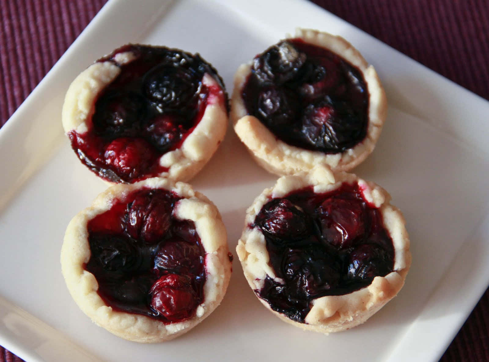 Blueberries Tart 桌布 壁紙 — 四個迷你莓果塔，帶有光澤的深色水果餡料，放在白色方形盤子上，背景為深紫色紋理。