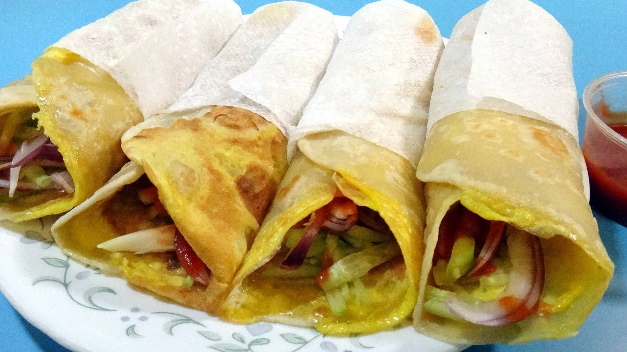 Egg 桌布 壁紙 — 四個金黃色的捲曲可麗餅，內有色彩繽紛的蔬菜和醬汁，放在一個花紋盤上，背景為藍色。